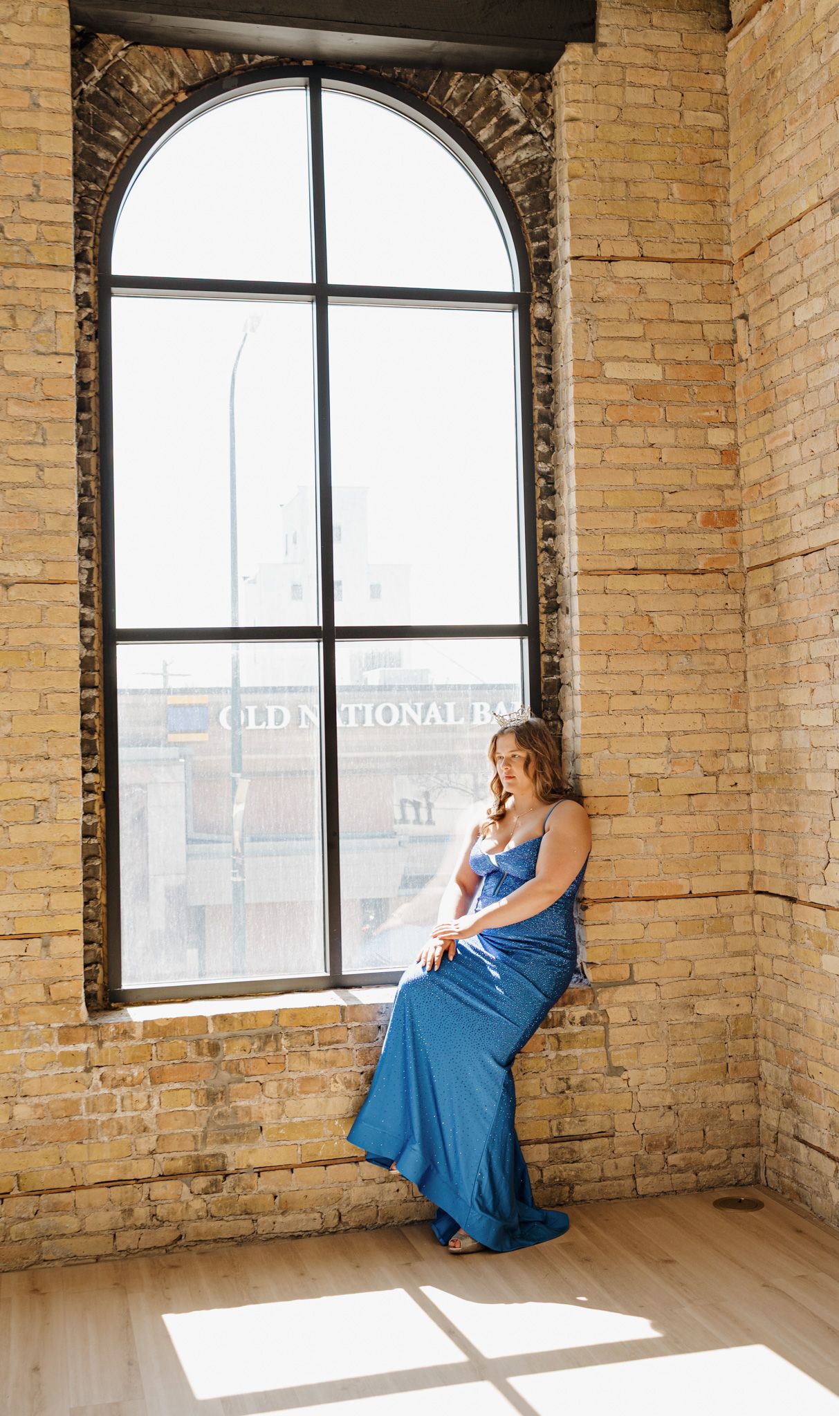 Woman in blue gown poses by arched window in brick loft near Old National Bank building.