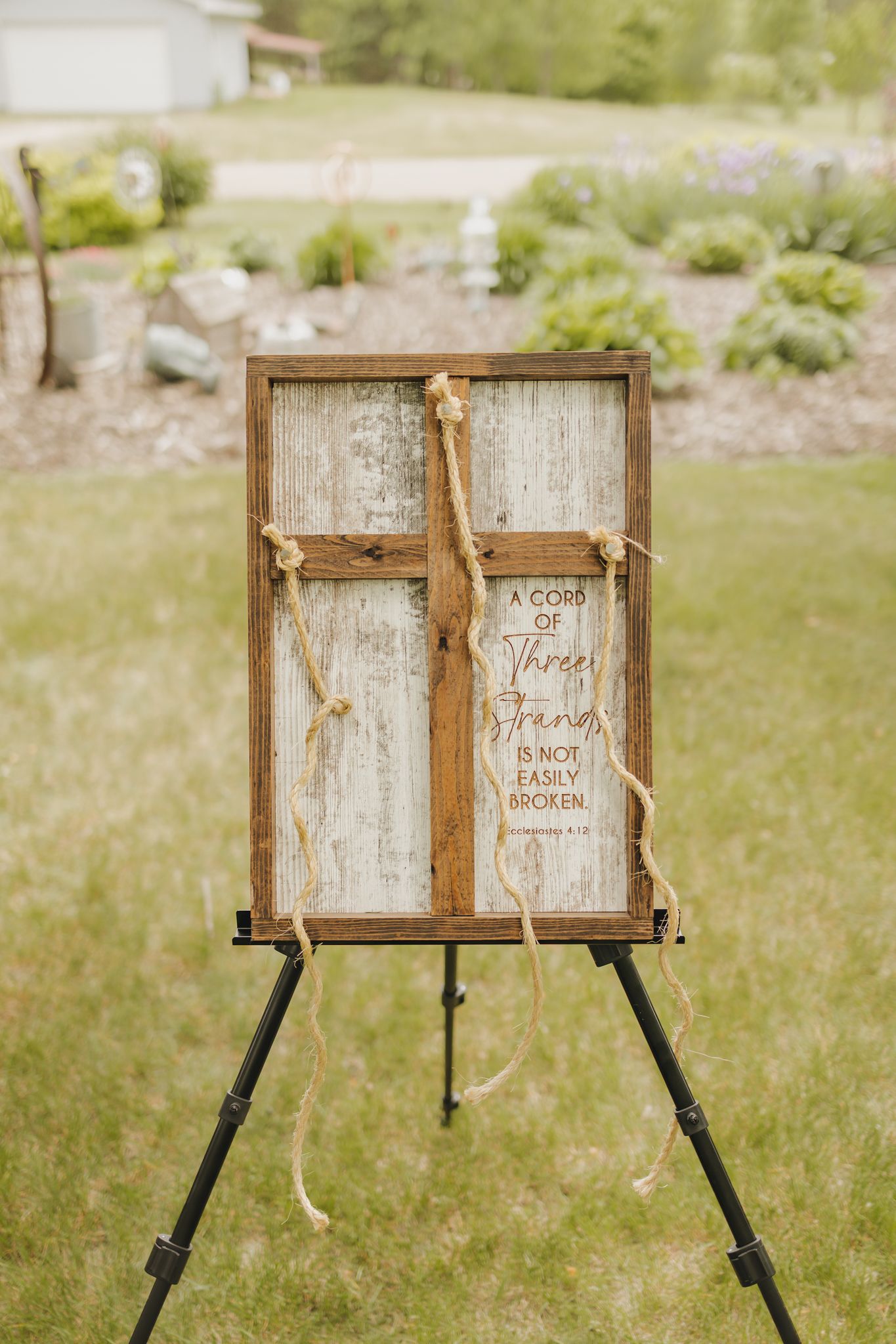 Rustic wooden wedding sign on easel reads A Cord of Three Strands is Not Easily Broken outdoors.