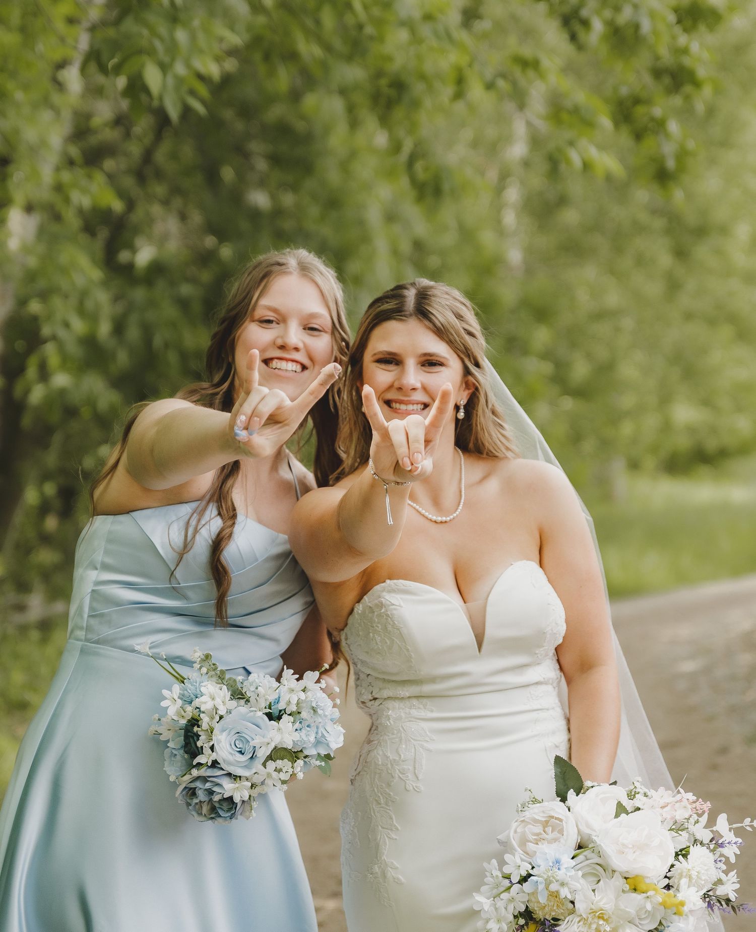 Bride and bridesmaid in blue dress posing with shaka signs holding floral bouquets outdoors.