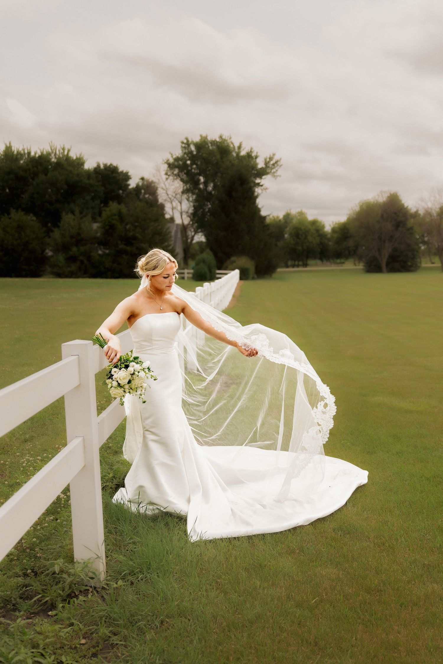 Bride in strapless white gown with flowing veil caught in wind, holding bouquet by white fence on green lawn.