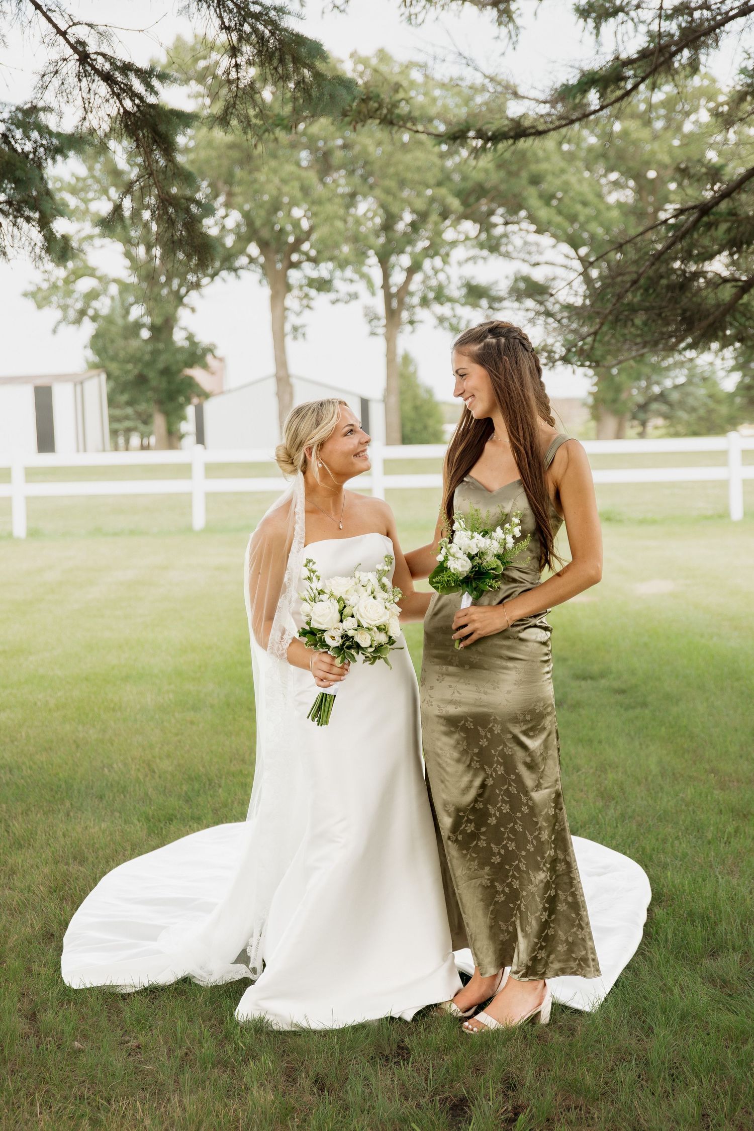 Bride in white gown and bridesmaid in olive dress holding bouquets laughing outdoors near white fence.