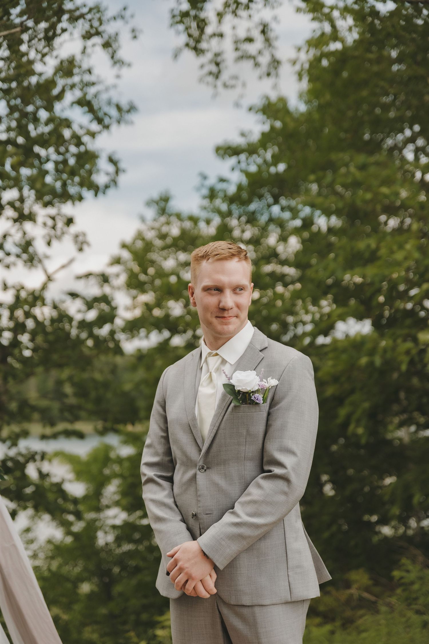 Smiling groom in gray suit with white boutonniere stands outdoors surrounded by lush green trees.