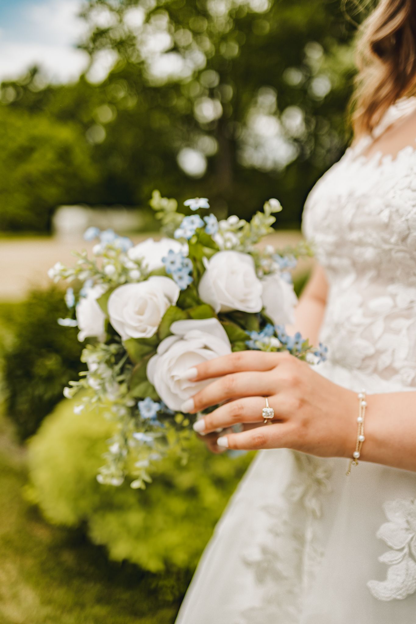 Bride holding white rose and blue flower wedding bouquet wearing diamond ring and gold bracelet in lace gown.