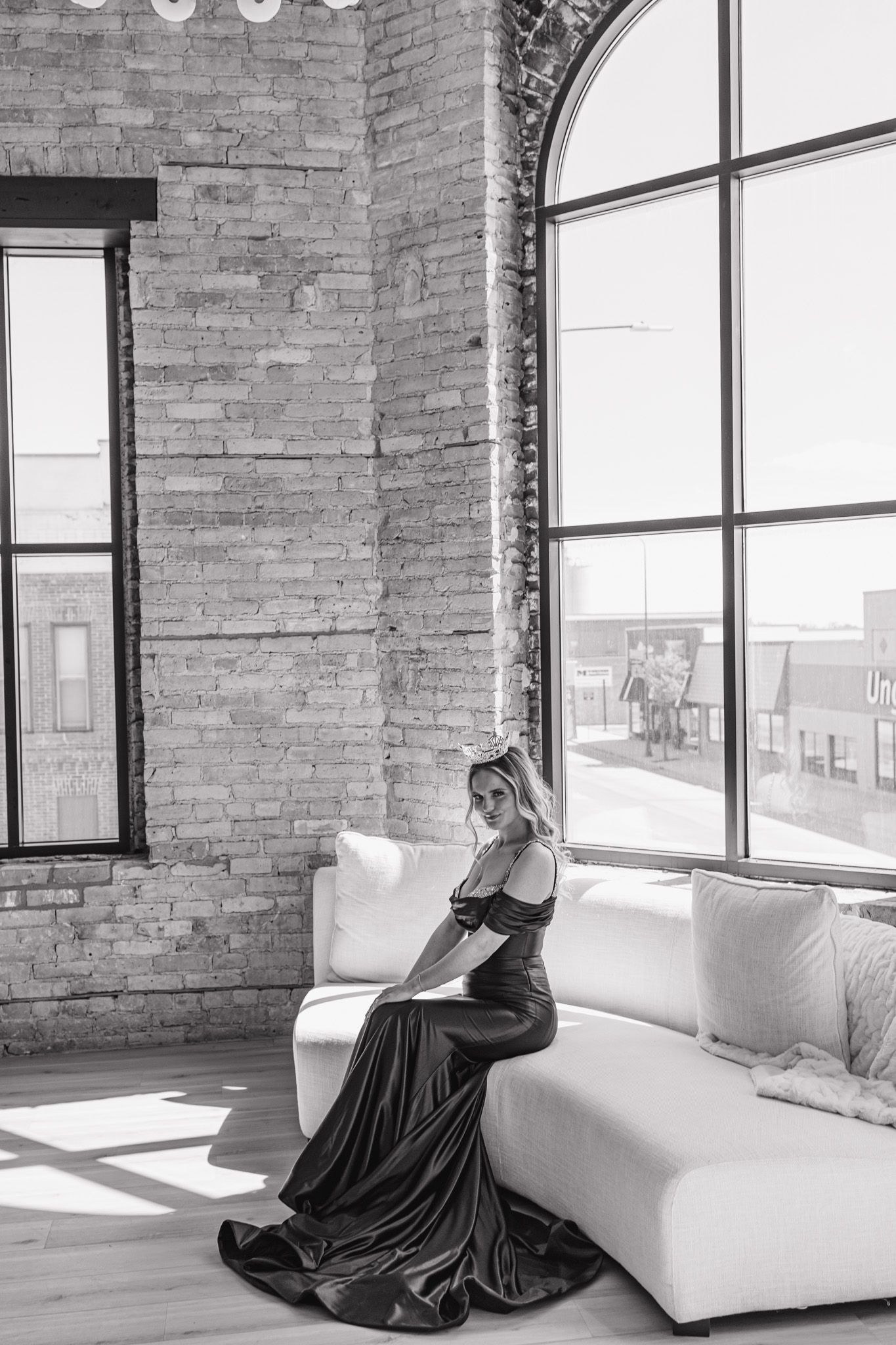 Woman in elegant dark gown seated on white sofa in industrial loft with brick walls and arched windows.