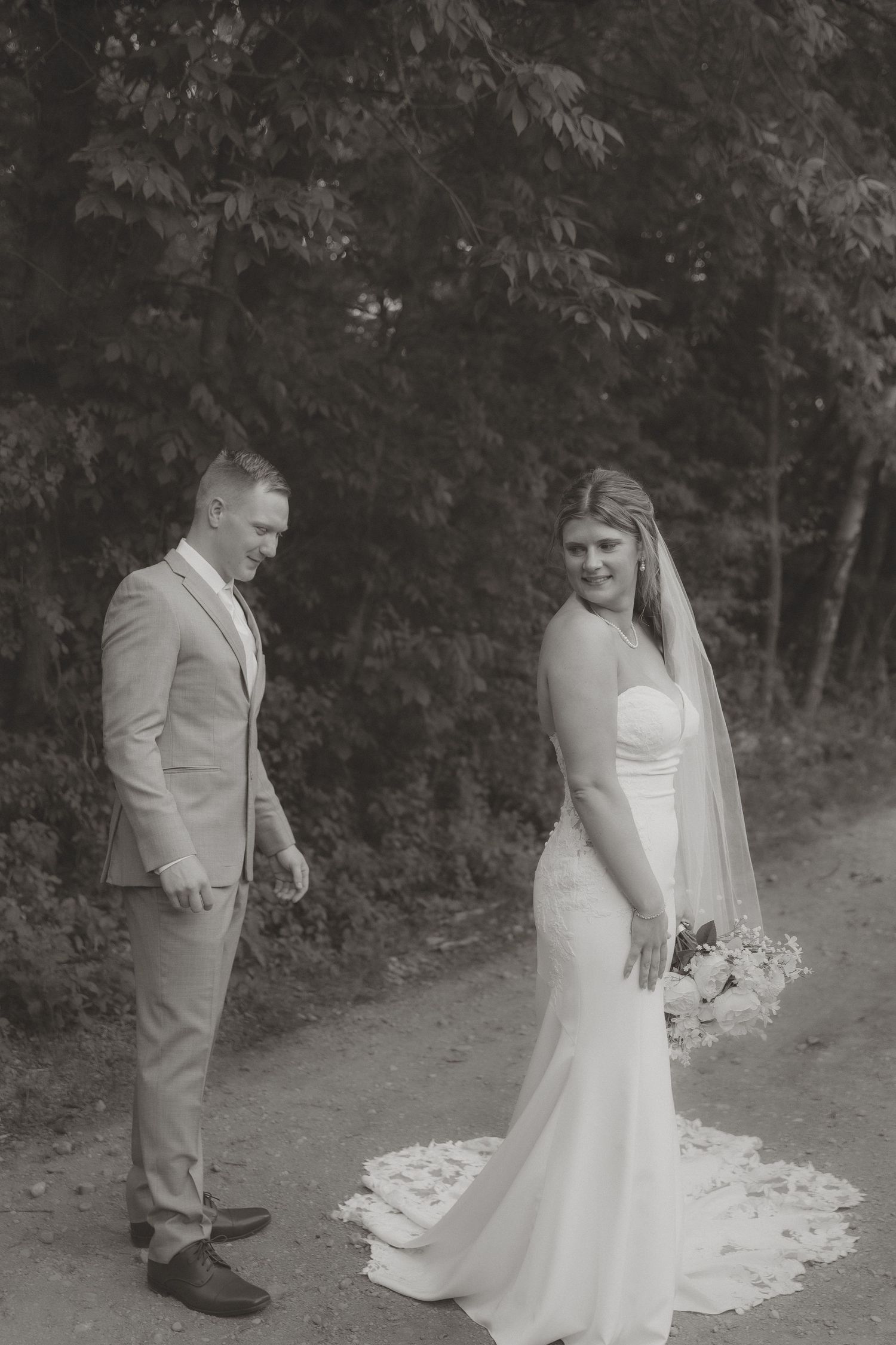 Black and white photo of bride in fitted gown with veil and groom in suit posing on a dirt path.