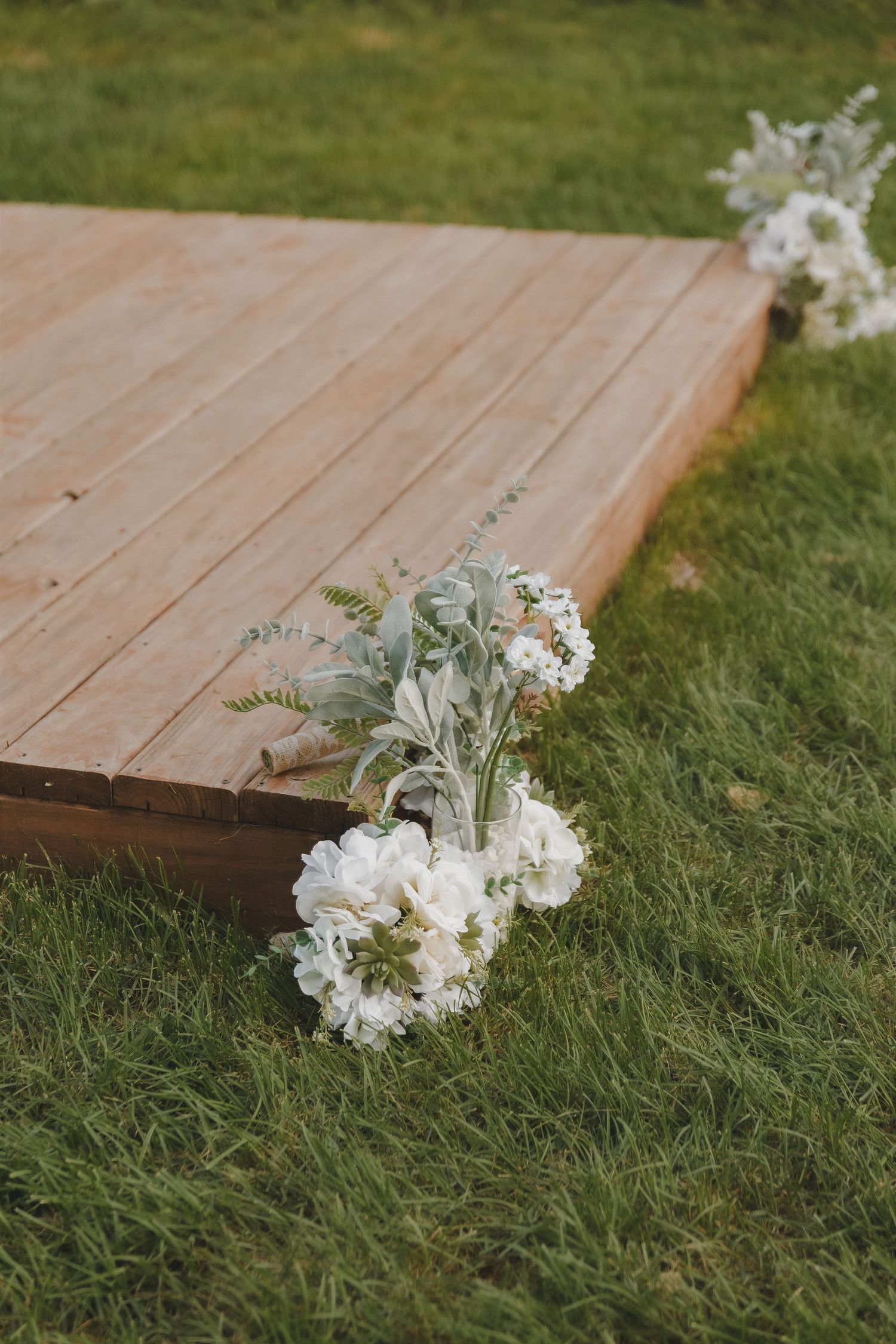 White floral wedding ceremony decorations placed along edge of wooden platform on green grass.