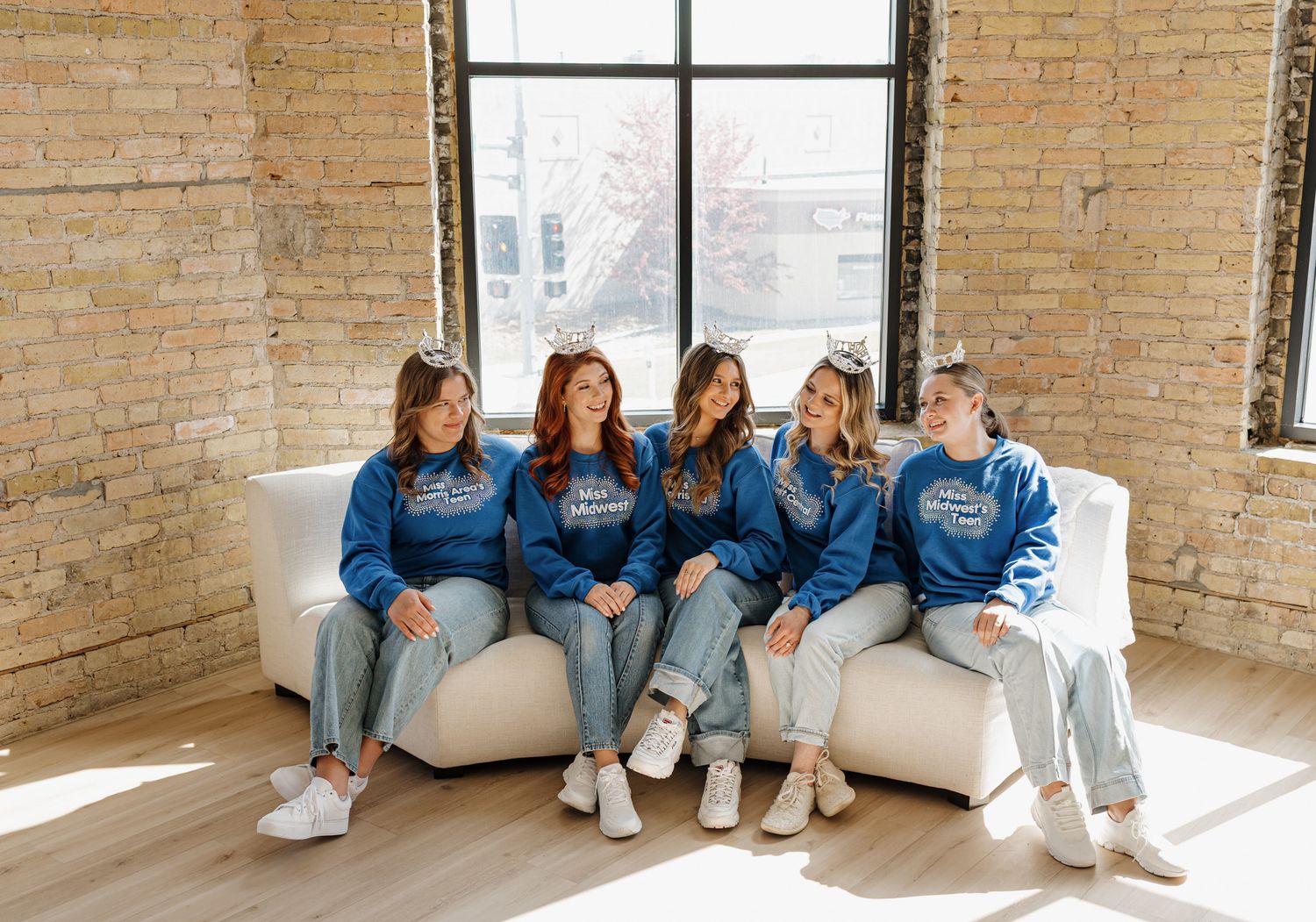 Five women in matching blue sweatshirts and jeans sit together on a white couch in a bright brick-walled room.