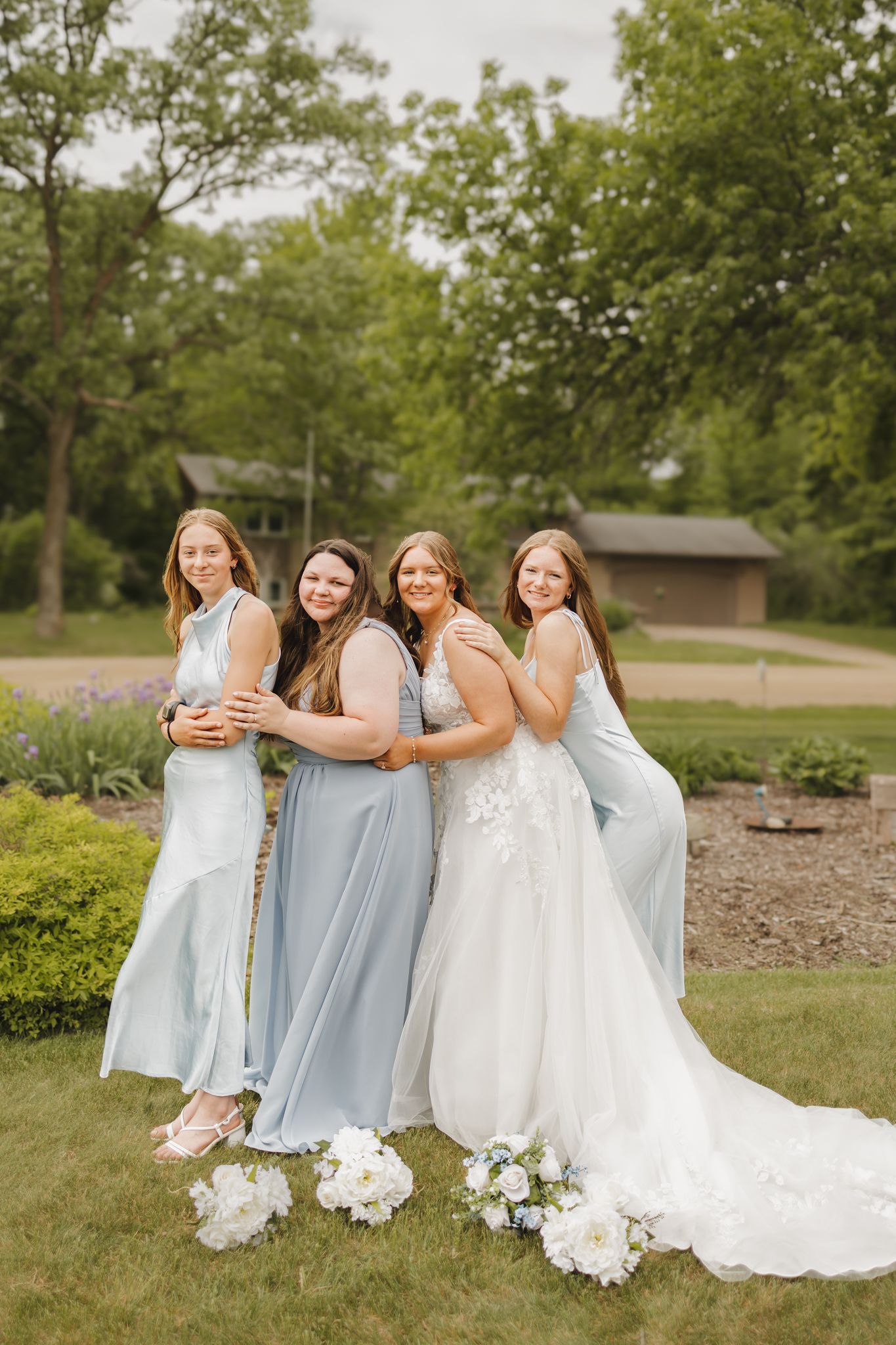 Bride in white lace gown poses with three bridesmaids in light blue dresses in garden setting.