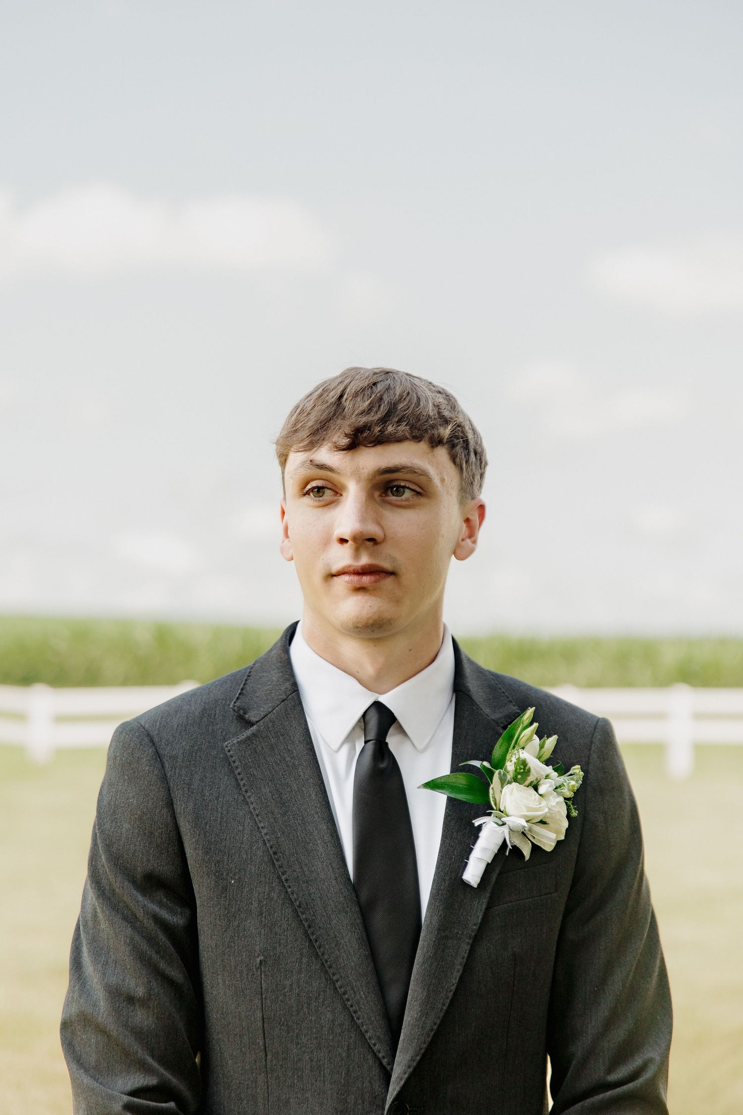 Young groom in dark gray suit with white boutonniere poses outdoors at wedding venue with white fence.