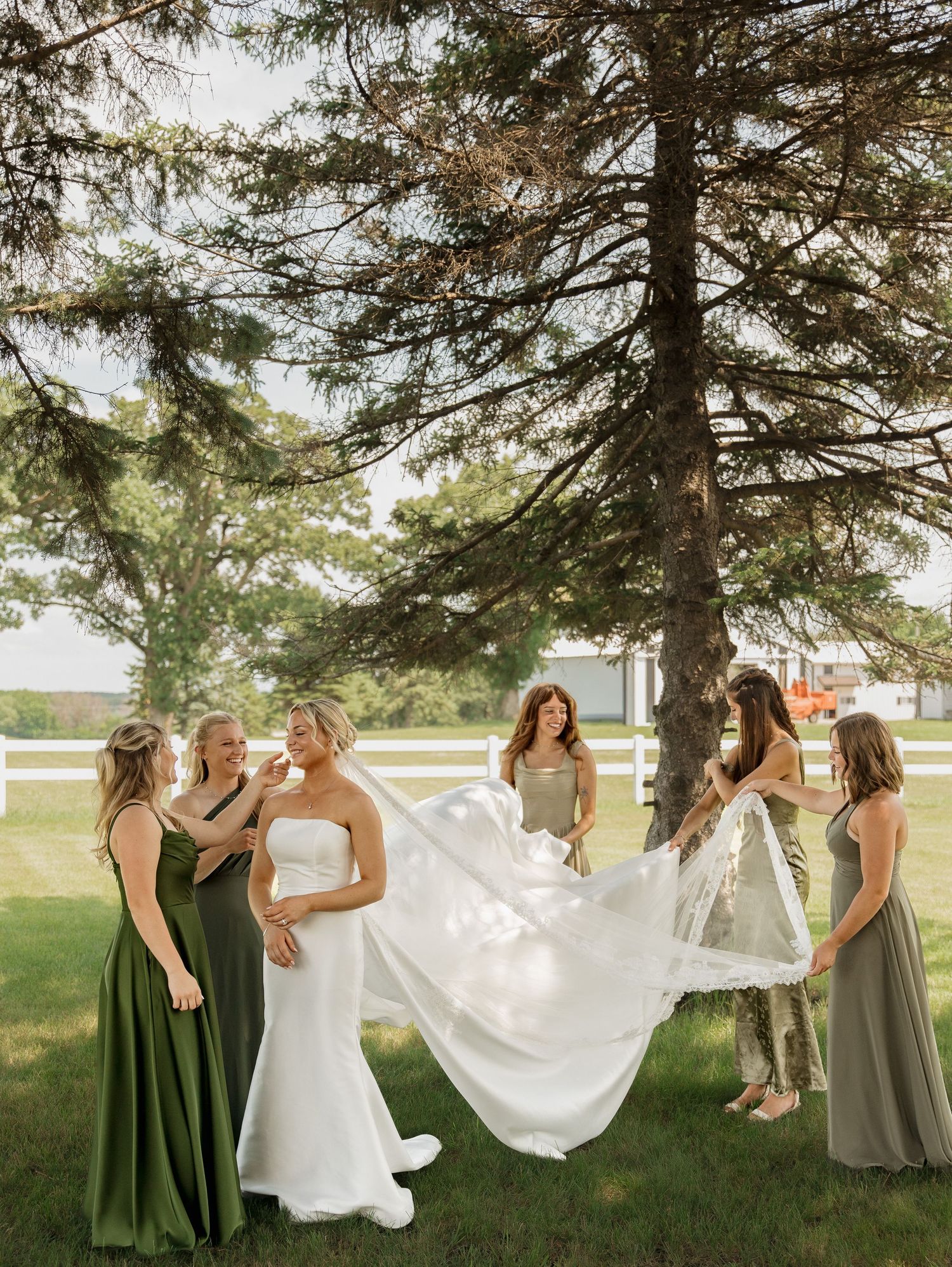 Bride in white strapless gown surrounded by bridesmaids in sage green dresses lifting her veil outdoors.