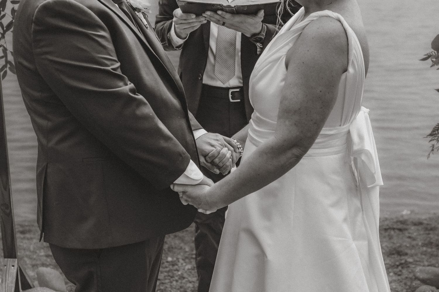 Bride and groom holding hands during wedding ceremony with officiant and lake backdrop.
