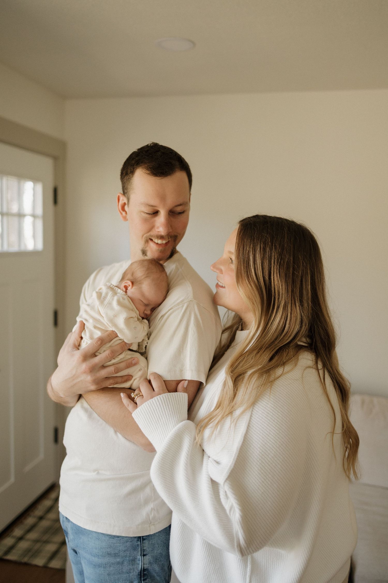 A smiling couple in white sweaters lovingly holds their newborn baby in a bright, cozy home.