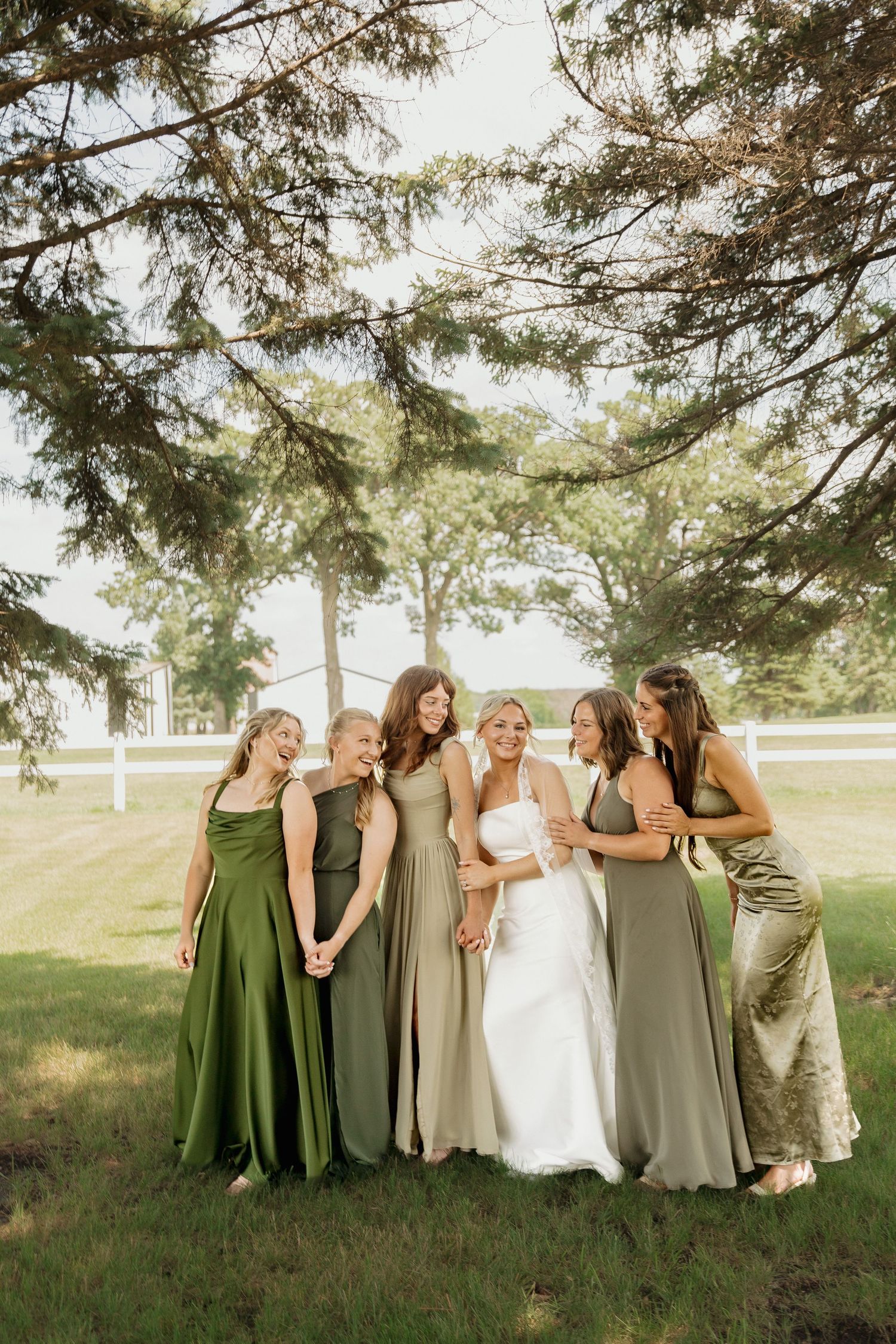 Bride in white gown surrounded by five bridesmaids in mixed green dresses laughing under pine trees outdoors.