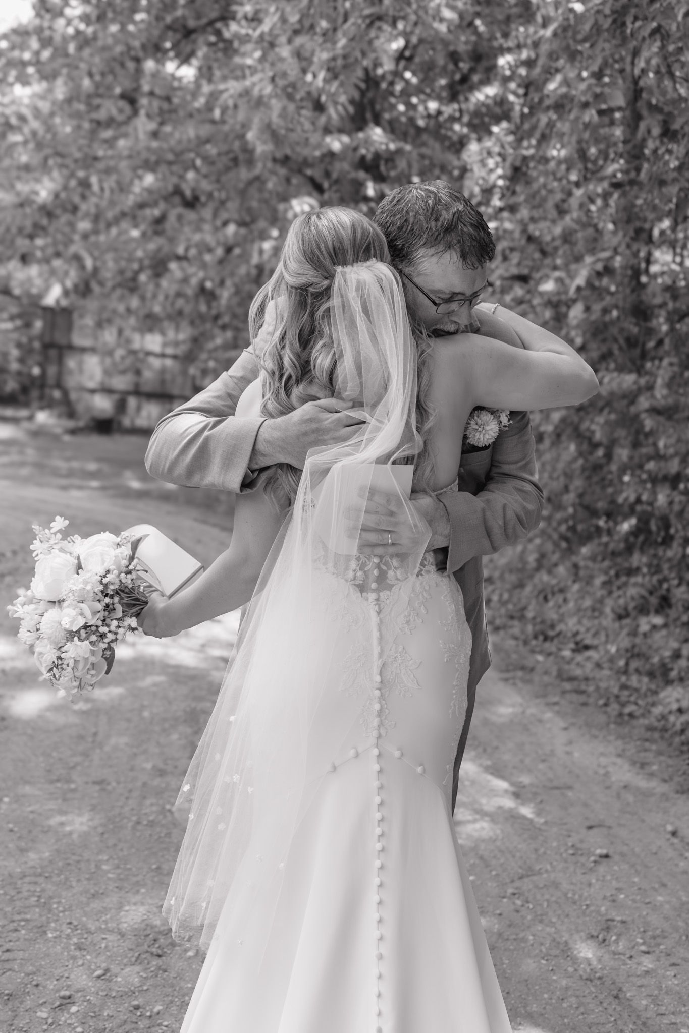 Black and white photo of bride and groom sharing an emotional hug outdoors, bride holding bouquet in wedding dress.