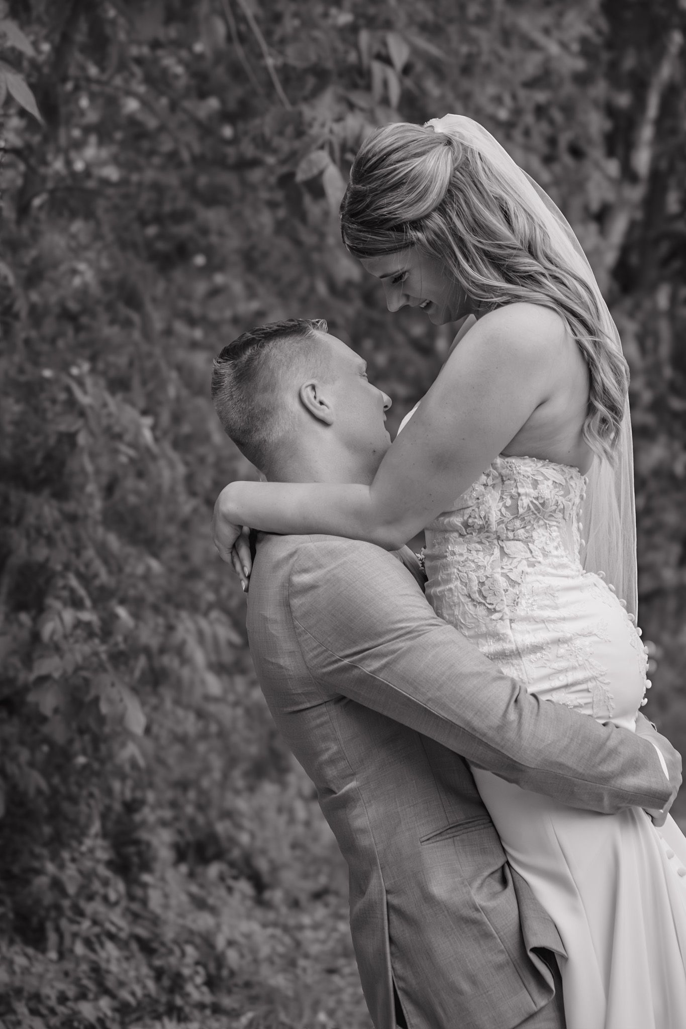 Black and white photo of groom lifting bride in wooded outdoor setting during wedding celebration.