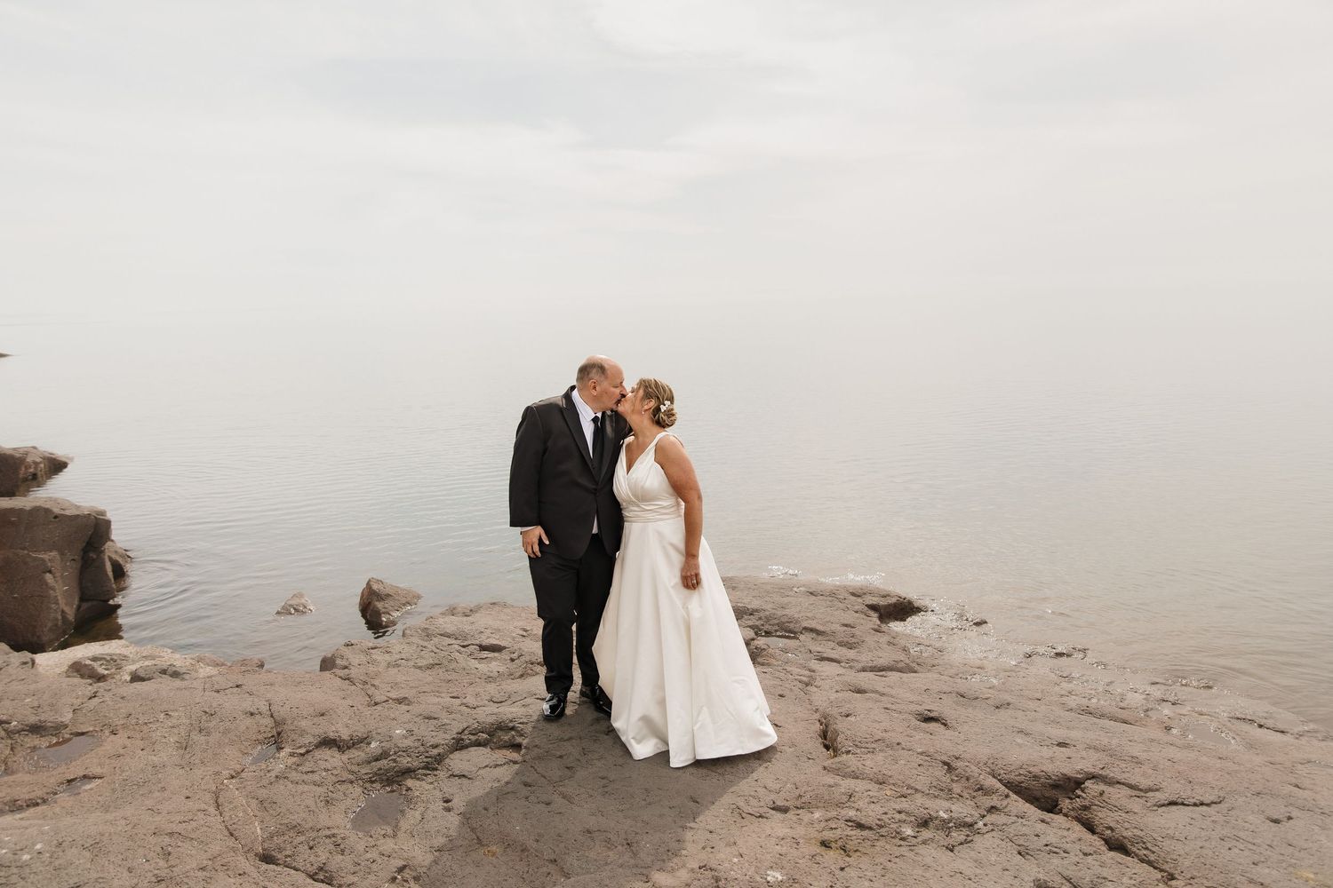 A bride and groom share a romantic kiss on rocky shoreline surrounded by misty fog on their wedding day.