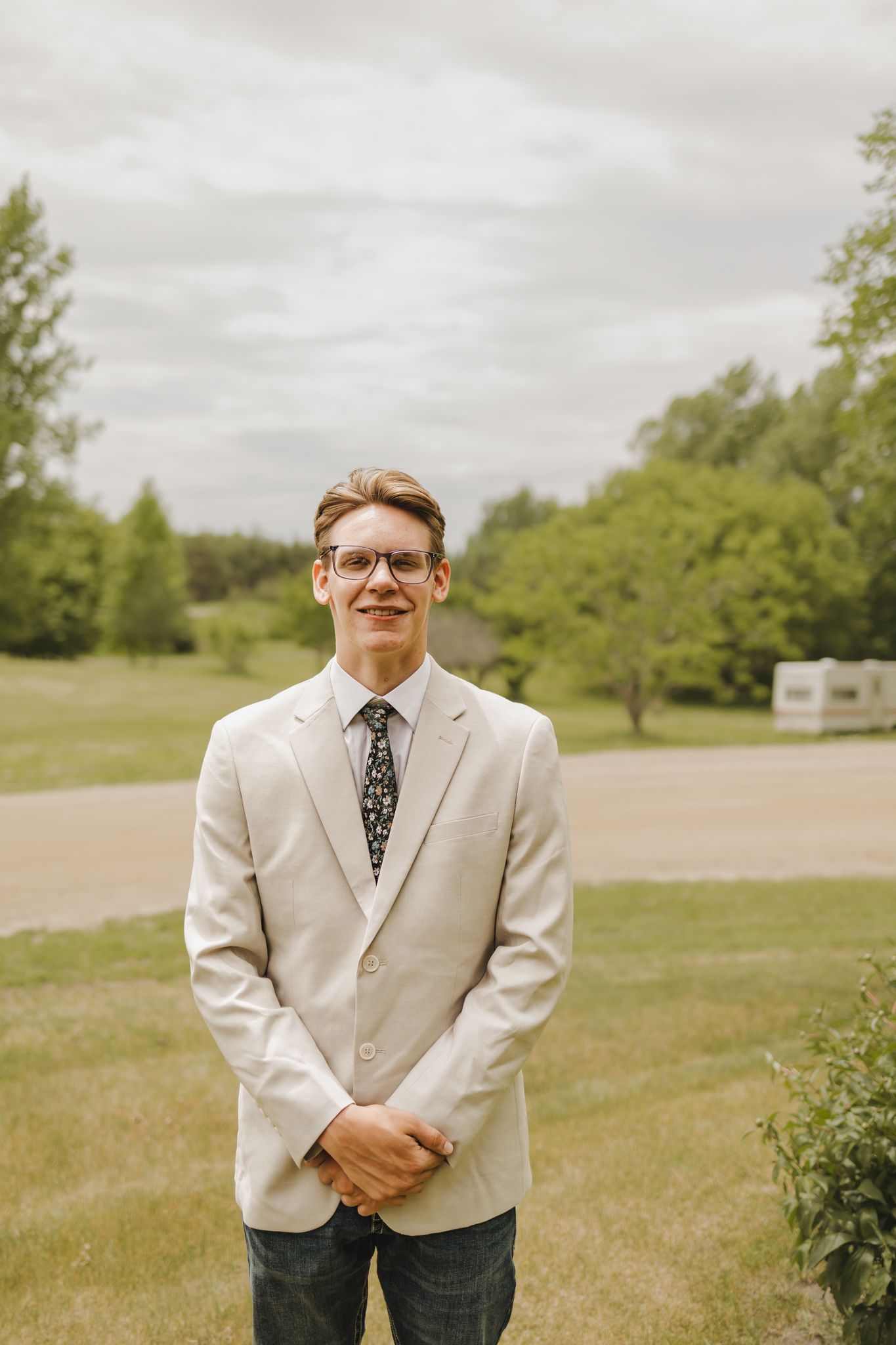 Smiling groom in a beige blazer and floral tie standing outdoors on a cloudy wedding day.