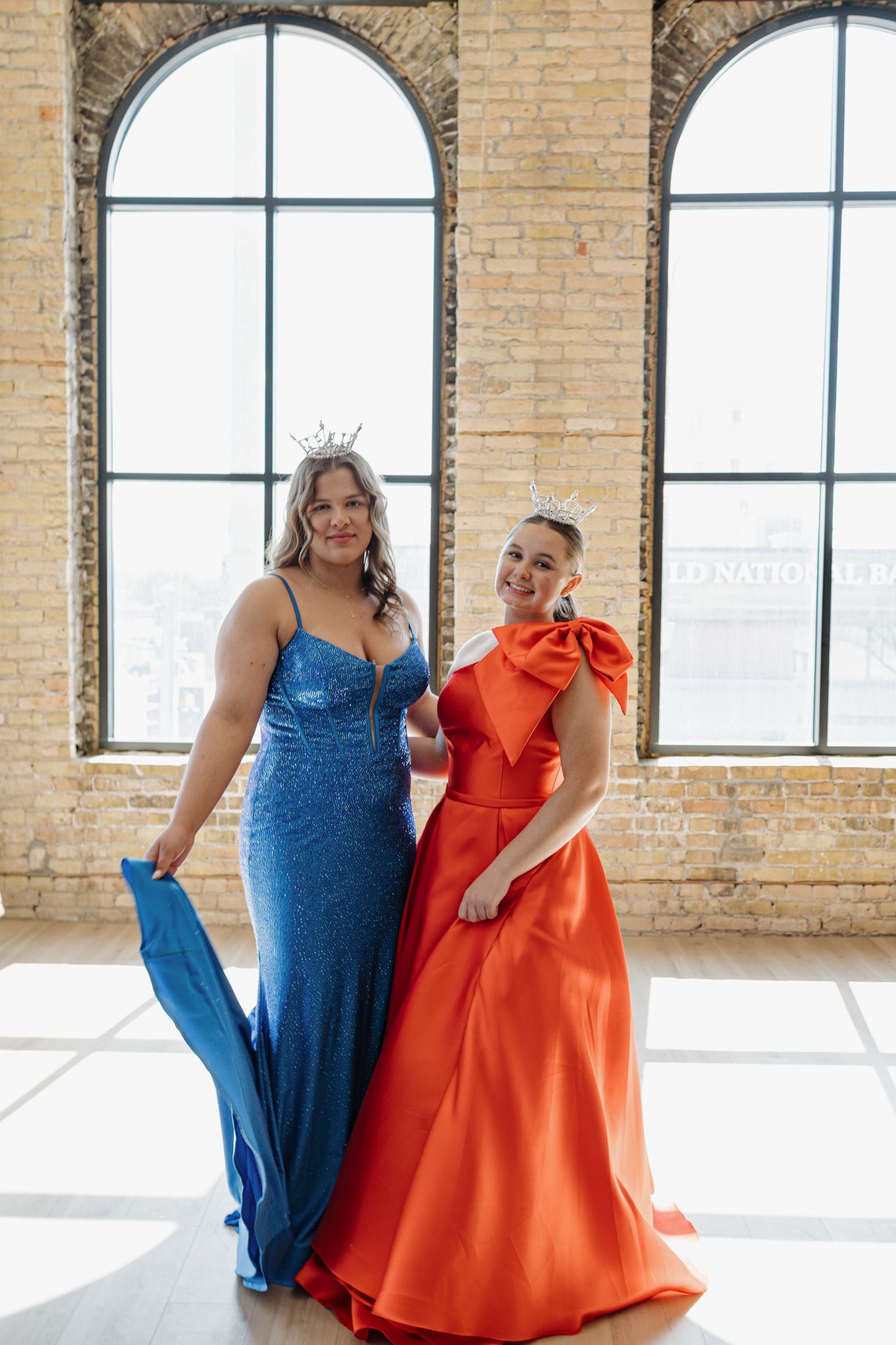 Two women in stunning formal gowns, one blue sequined and one orange bow dress, pose by arched windows.