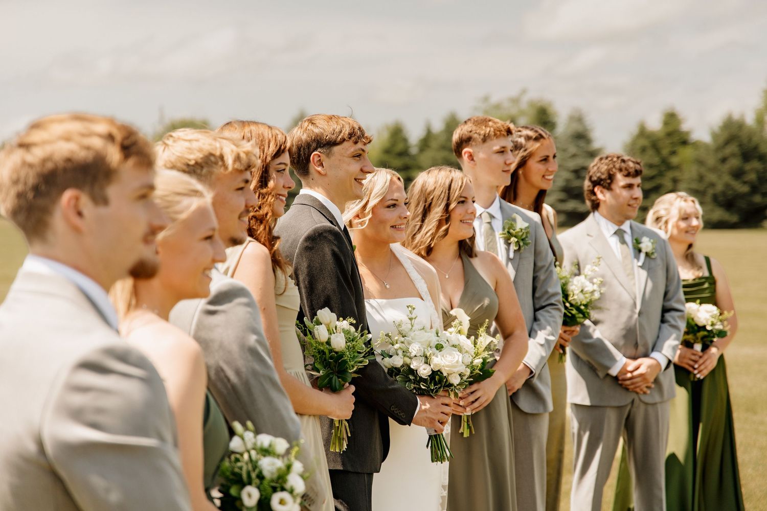 Wedding party in sage green attire holding white bouquets standing outdoors on a sunny day.