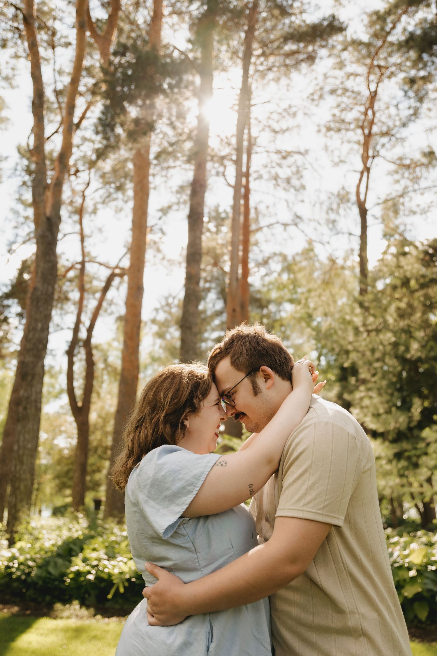 A couple embracing and touching foreheads in a sunny forest park, sharing a romantic moment outdoors.