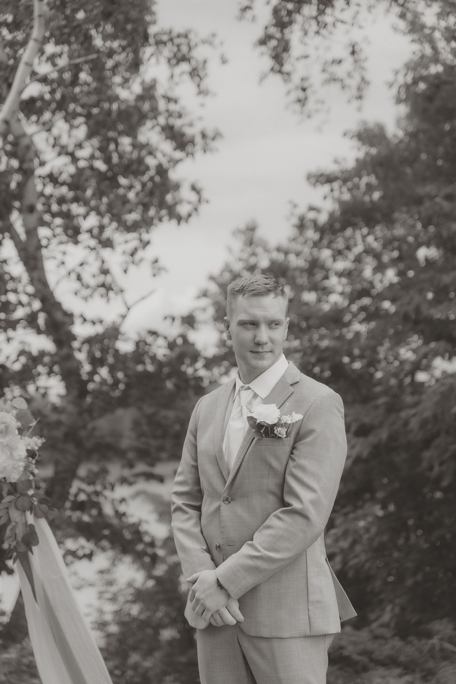 Black and white portrait of a groom in a gray suit with boutonniere standing outdoors at a wedding ceremony.