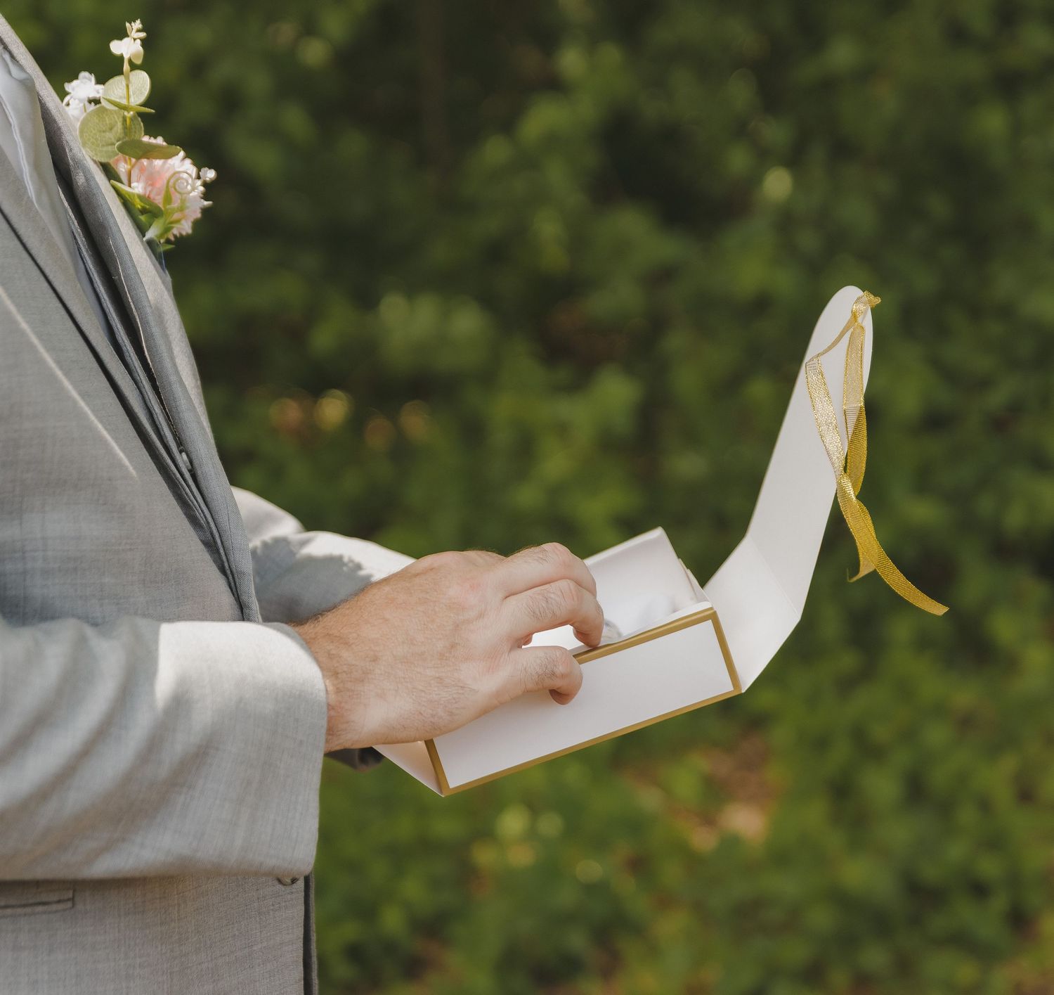 Groom in gray suit reads vows from gold-trimmed booklet during outdoor wedding ceremony.
