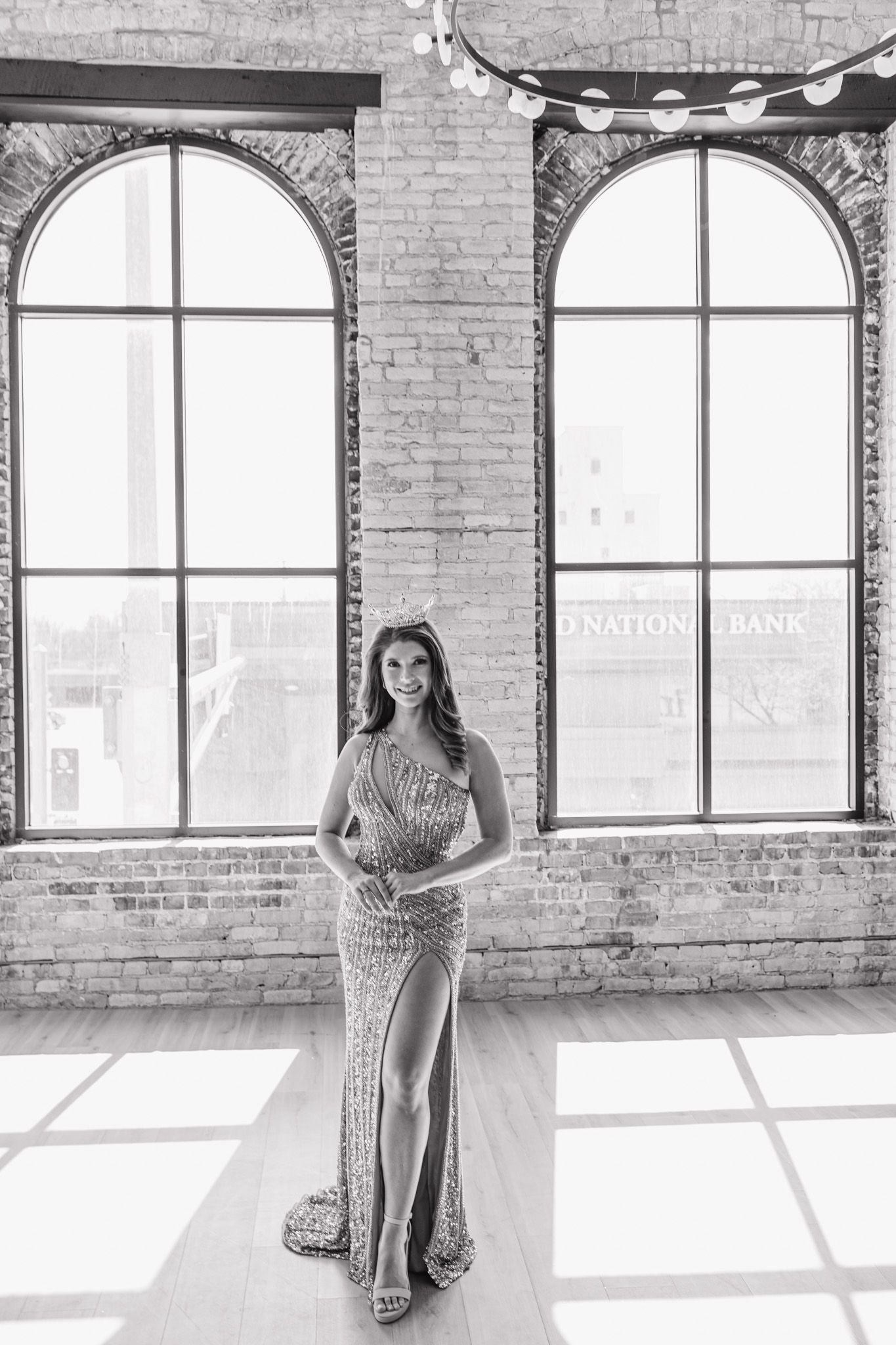 Black and white portrait of woman in sequin one-shoulder gown with crown in brick loft.