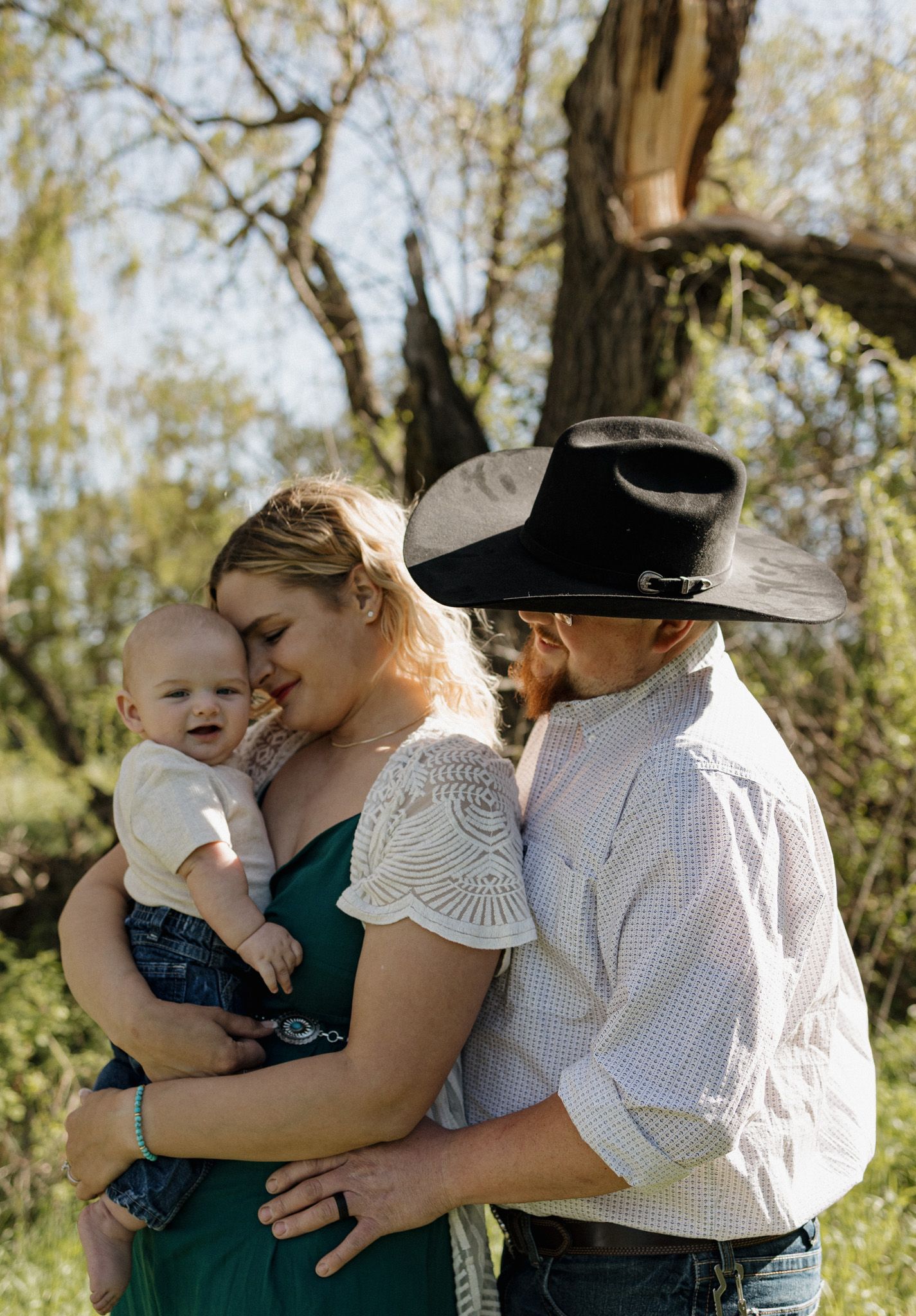A couple in a green dress and cowboy hat share a tender moment with their smiling baby outdoors.