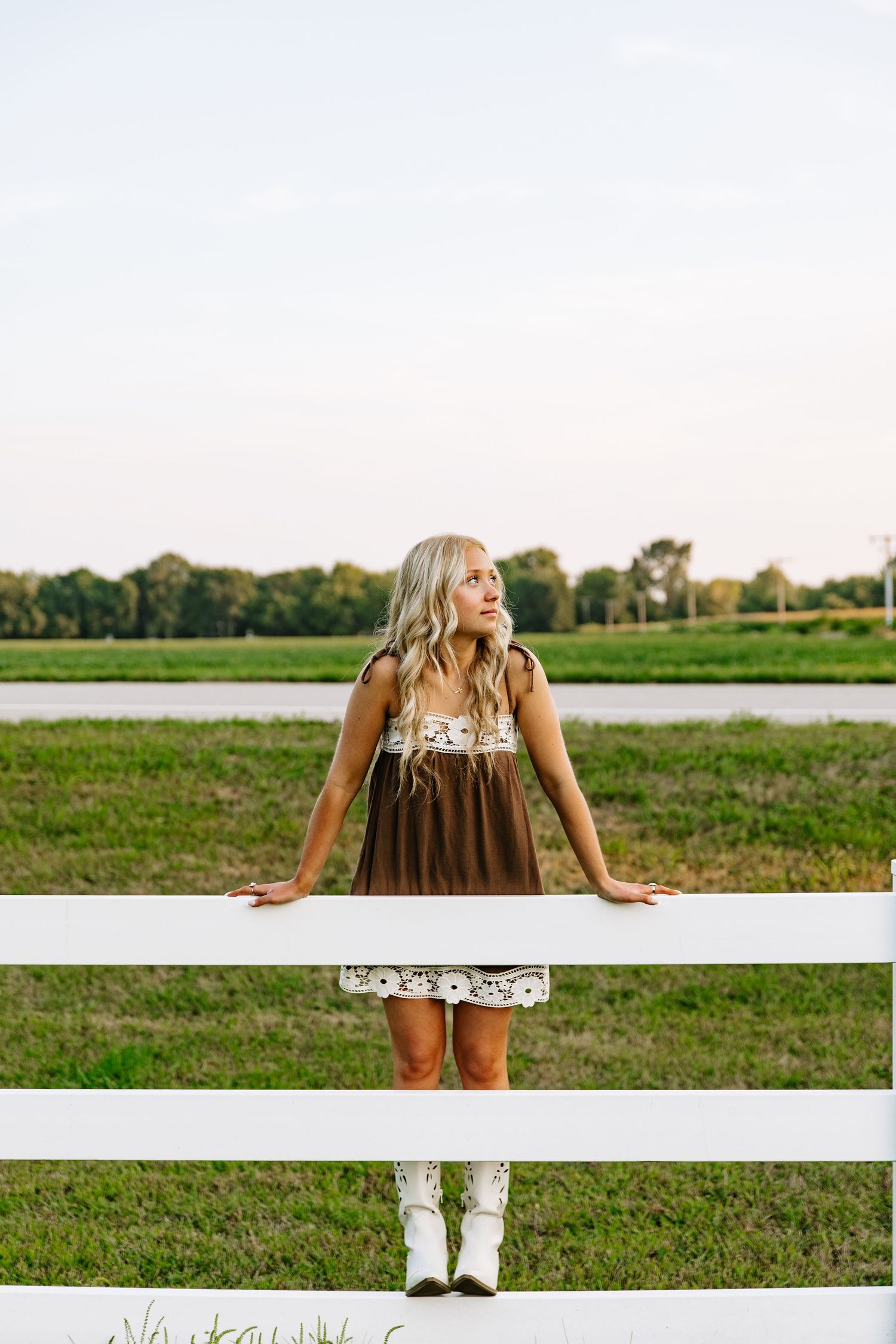 Blonde woman in brown dress and white boots leaning on a white fence in a green rural field.