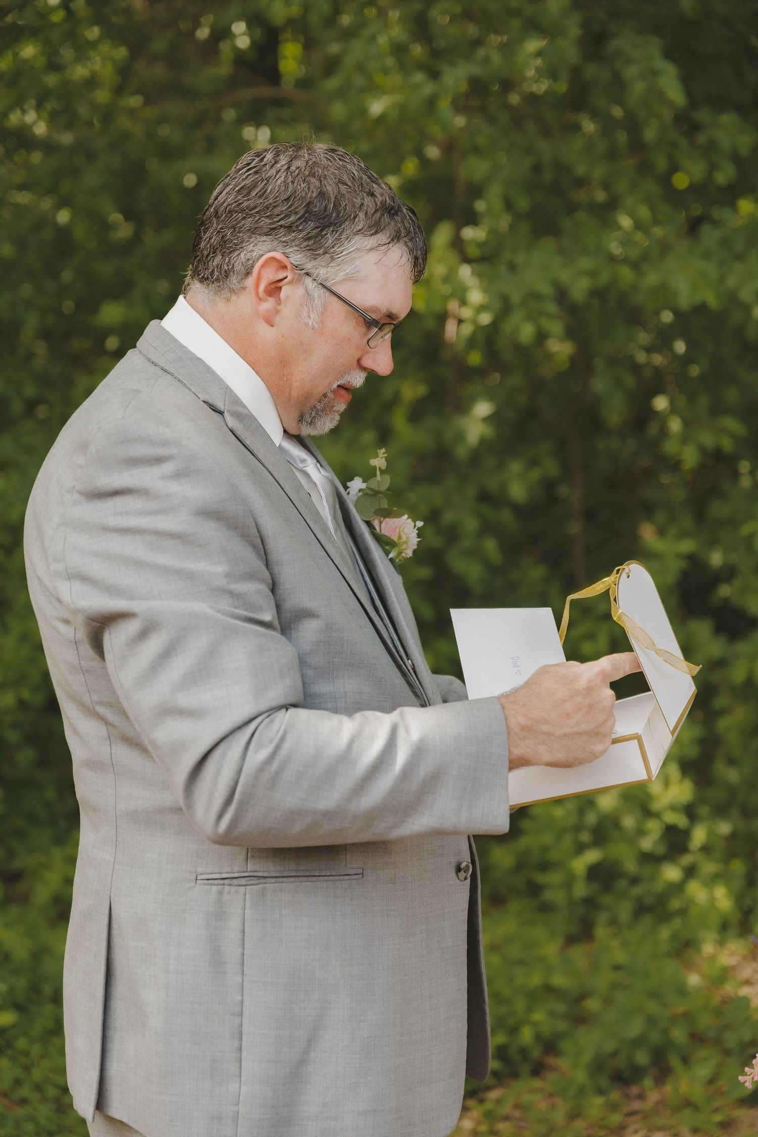 Groom in gray suit reads heartfelt wedding vows from a card outdoors surrounded by lush green trees.