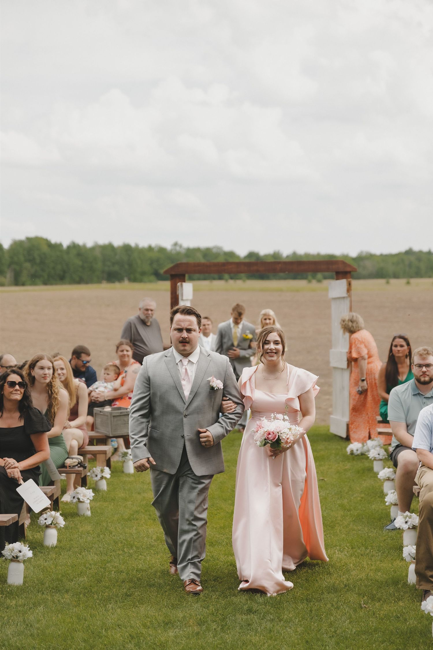 Groomsman escorts bridesmaid in pink dress down outdoor wedding aisle lined with white floral arrangements.