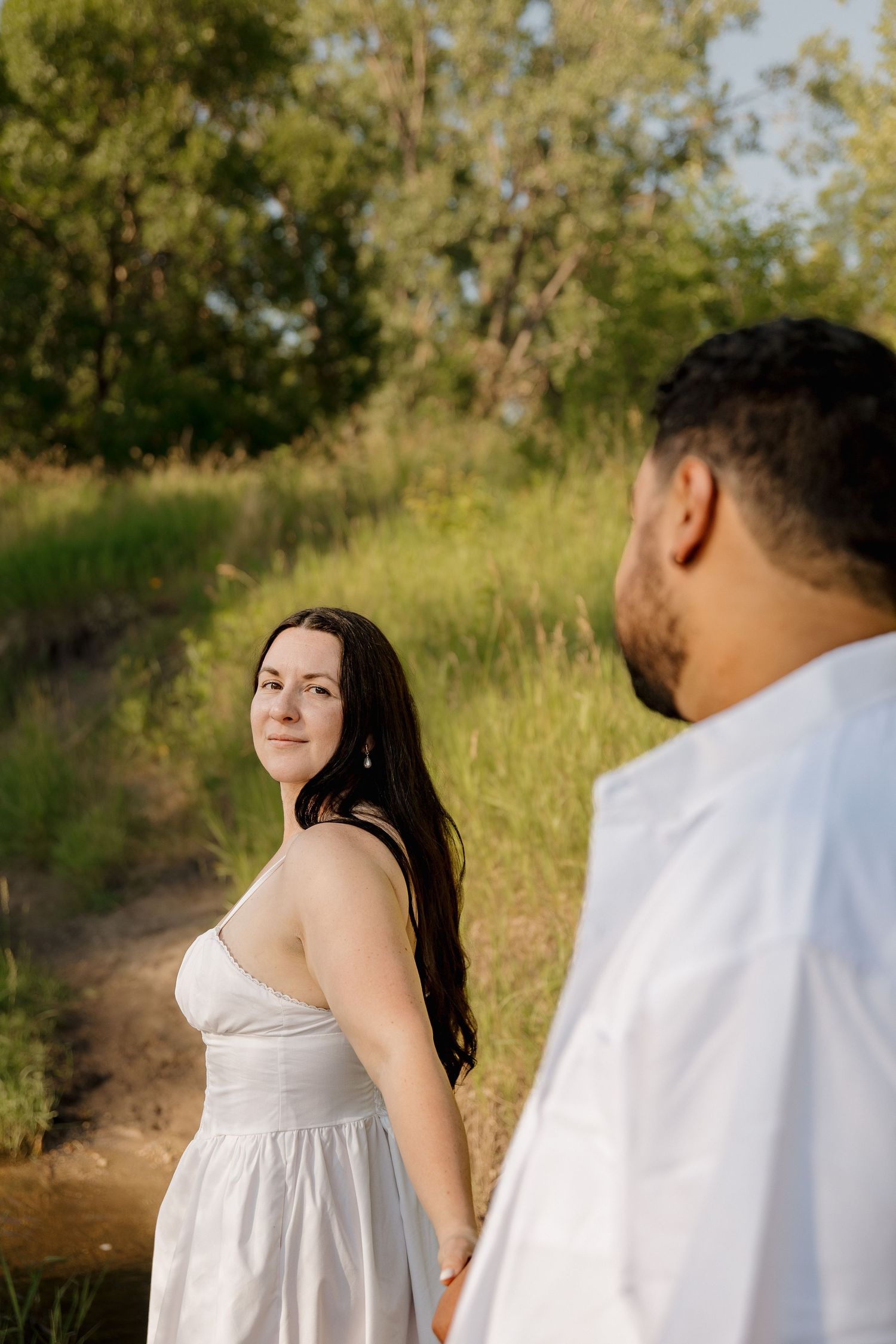 A woman in a white dress looks back over her shoulder at a man in white, standing in a sunny green meadow.