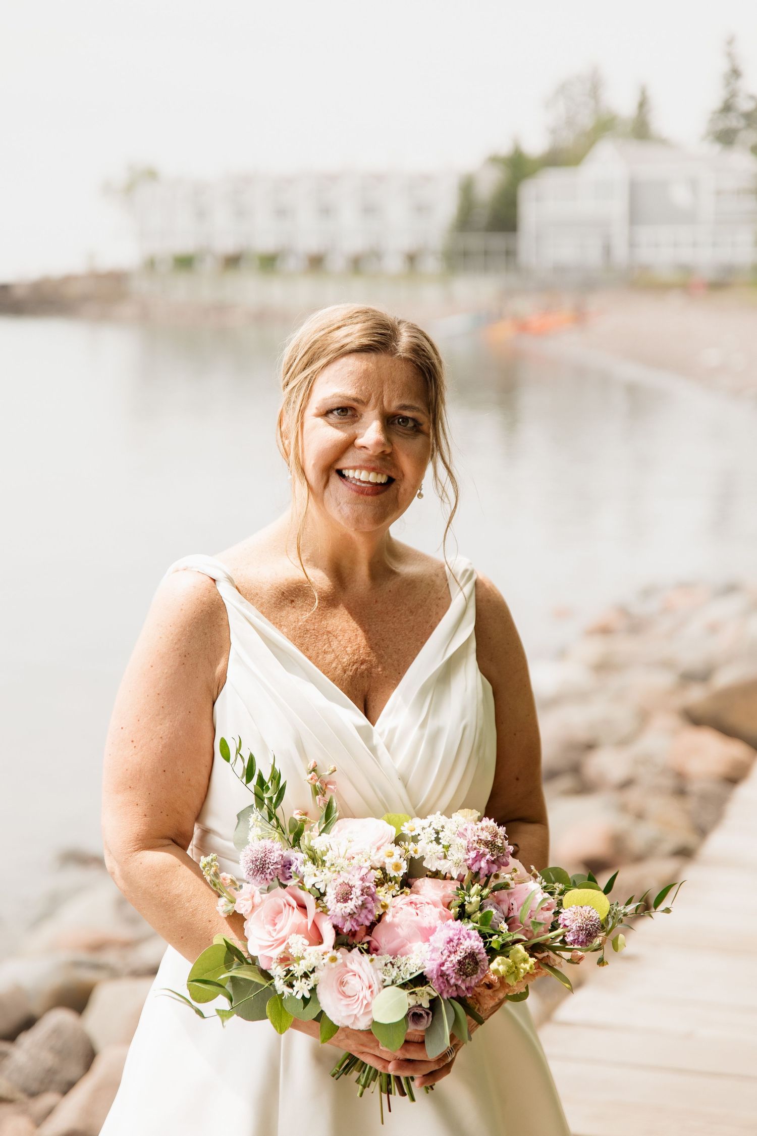 Smiling bride in white V-neck gown holding pink and purple bouquet by waterfront rocks.