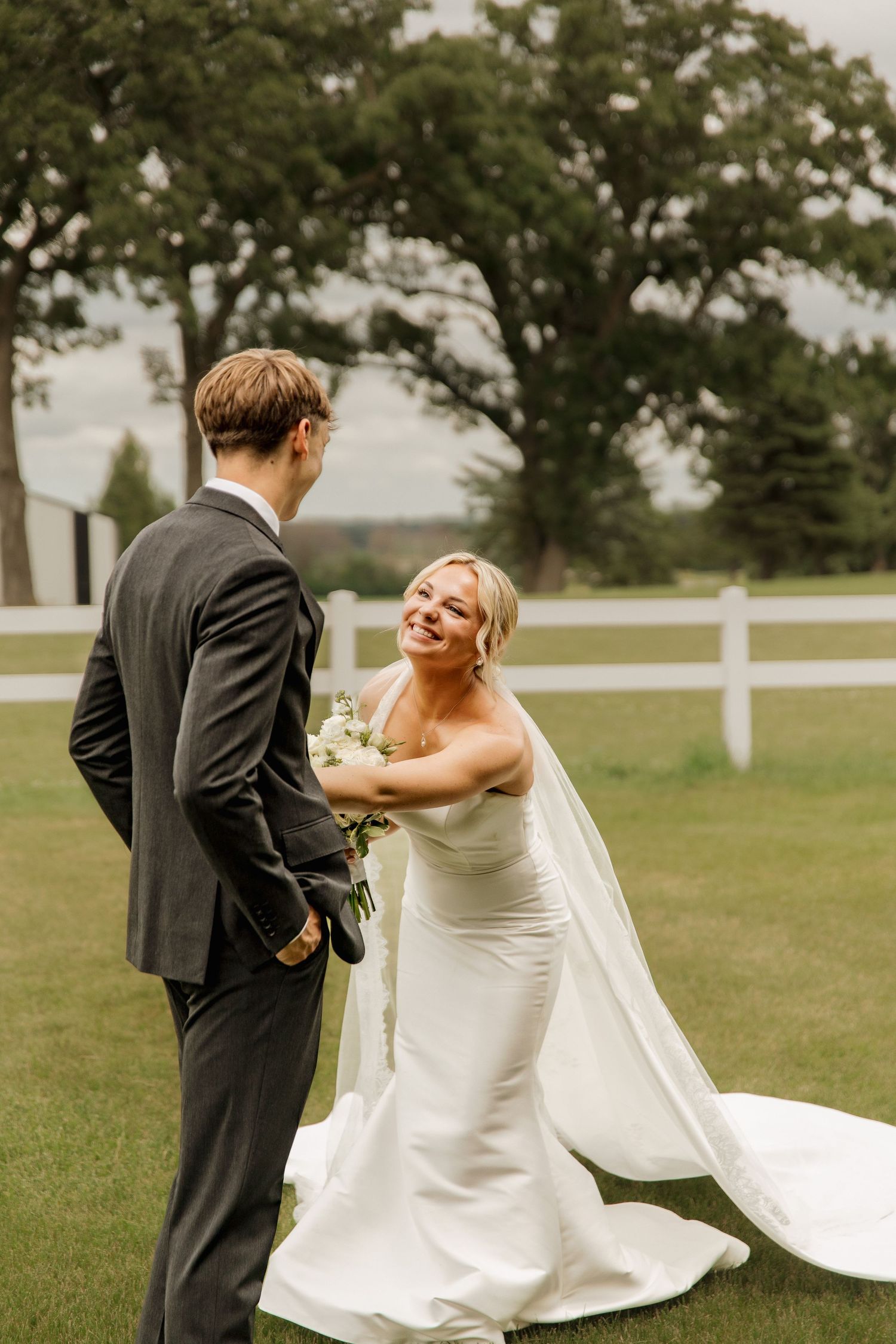 Smiling bride in strapless gown holding bouquet faces groom near white fence at outdoor farm wedding venue.