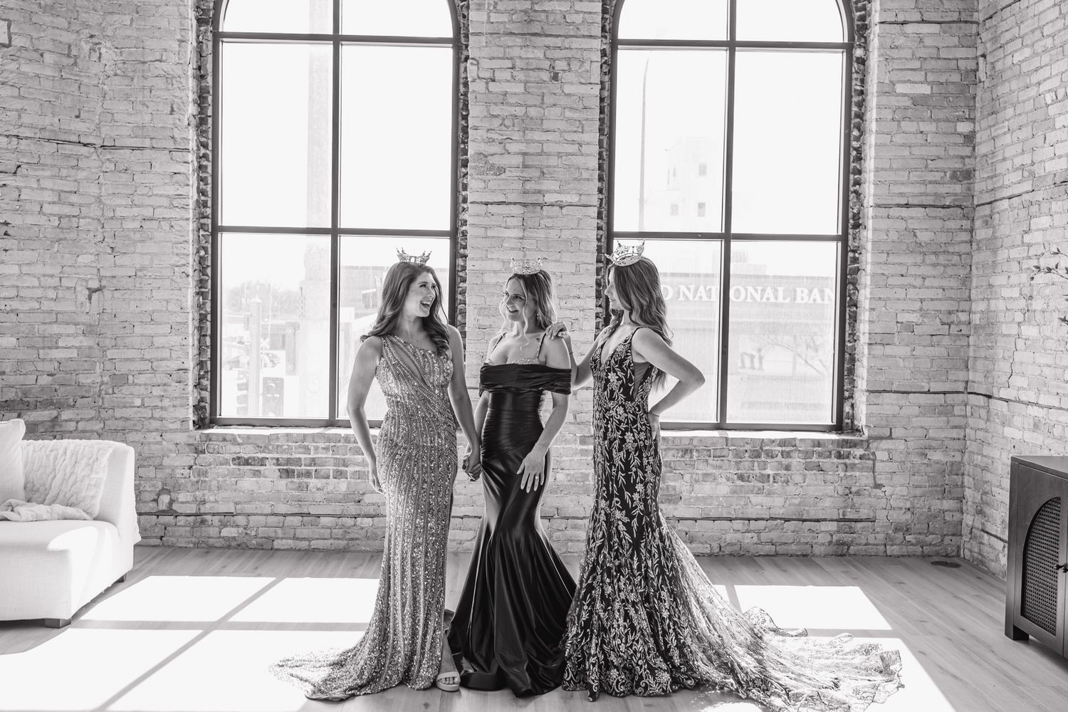 Three elegant women in formal gowns pose together in a black and white brick loft photo.