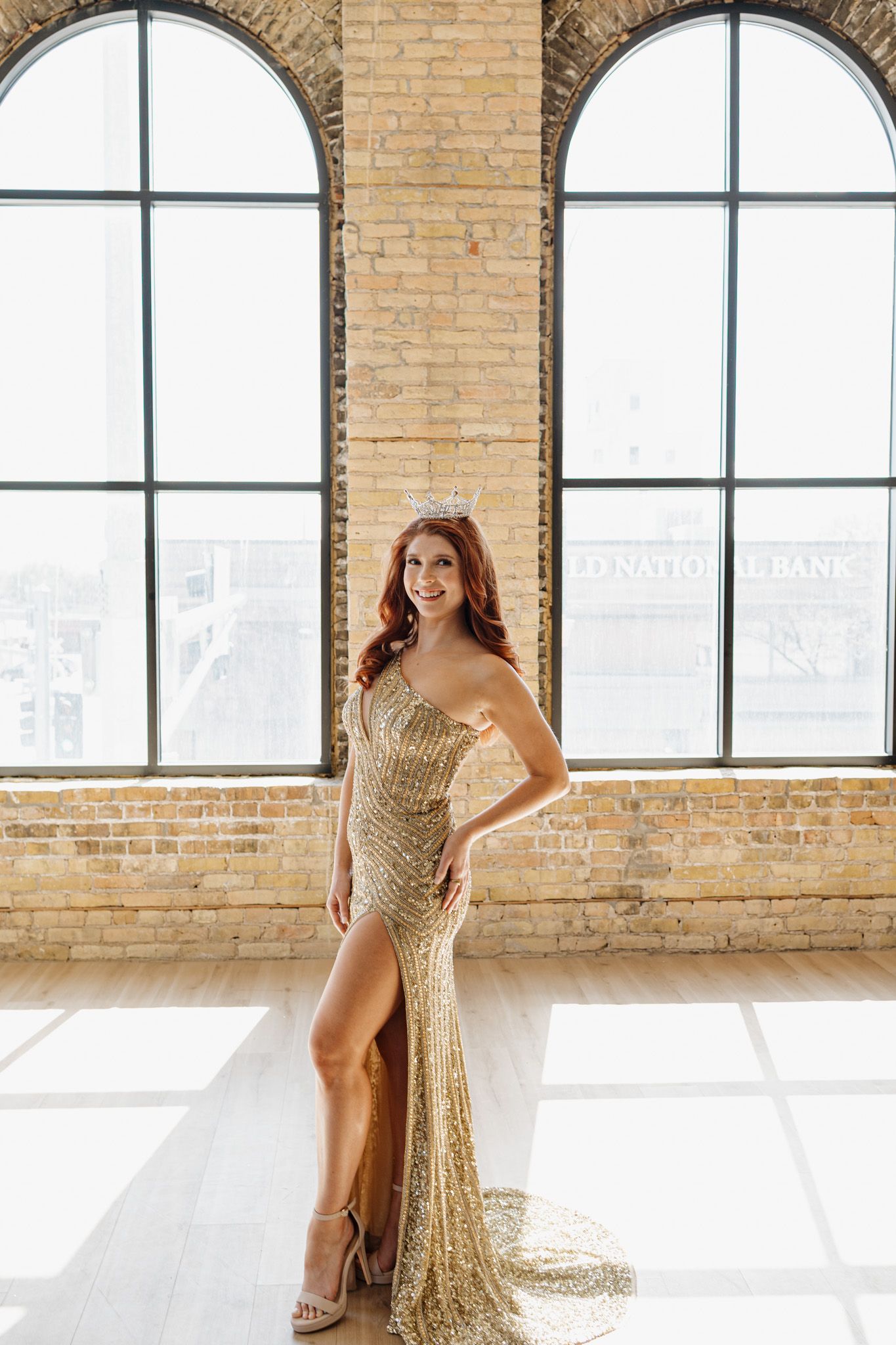 Stunning woman in gold sequin gown and crown poses in a bright brick-walled loft.