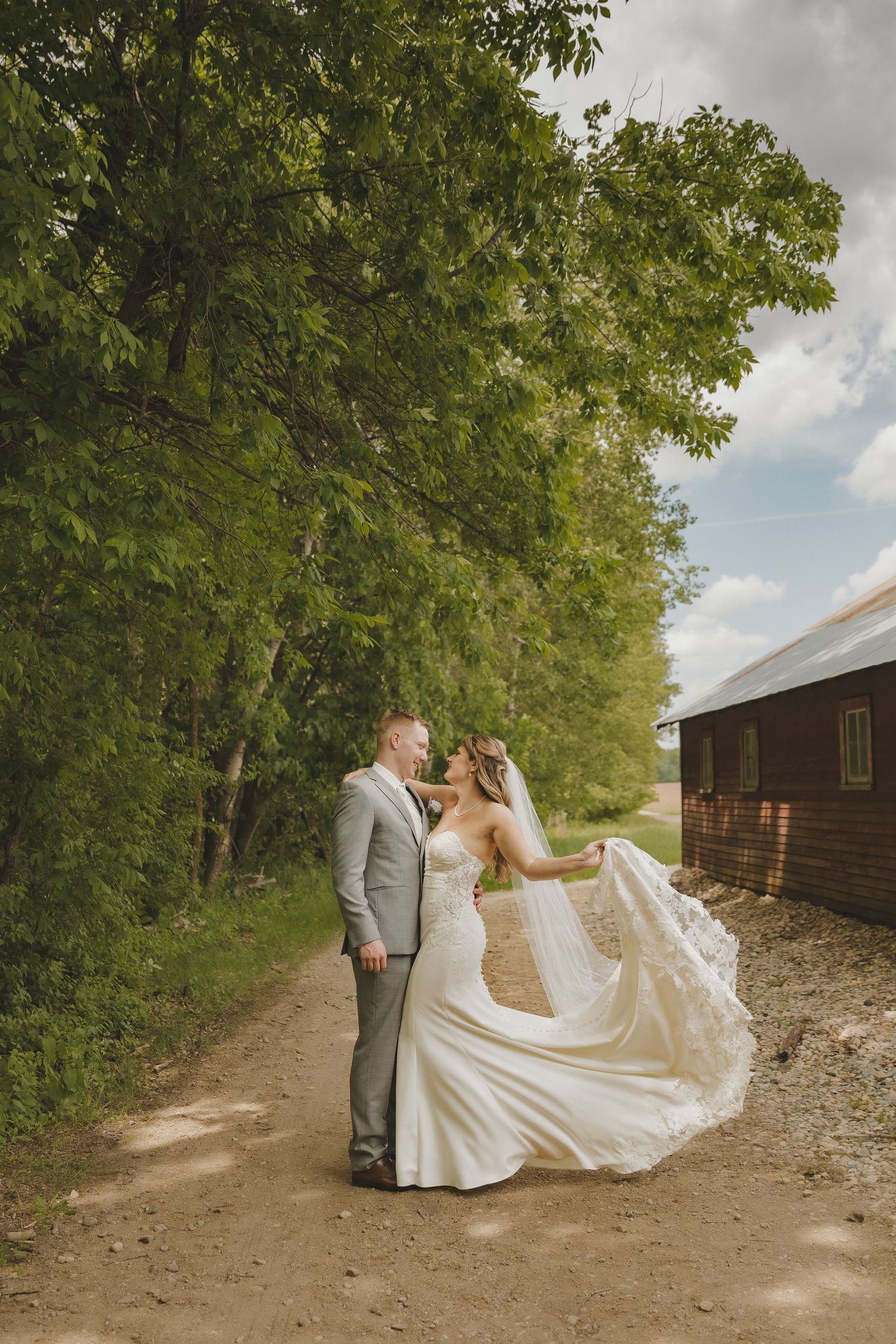 Bride and groom share a romantic moment on a rustic dirt path, her veil flowing in the wind, surrounded by lush green trees.