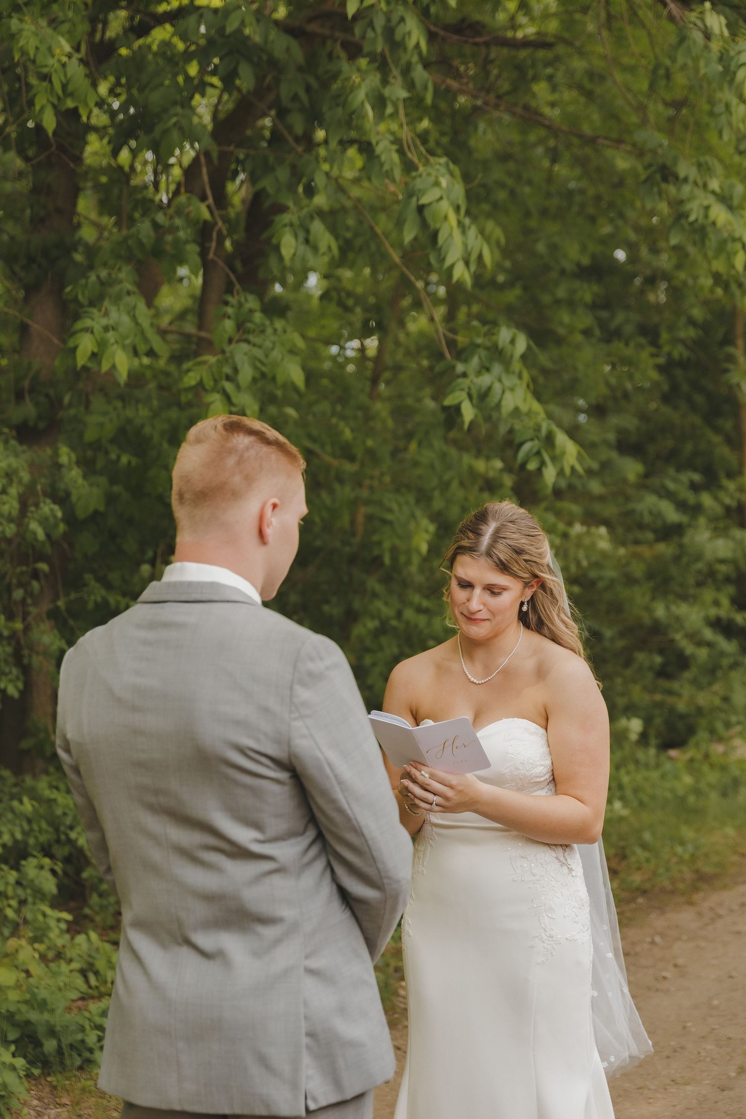 Bride in strapless white gown reads vows to groom in gray suit during outdoor woodland ceremony.