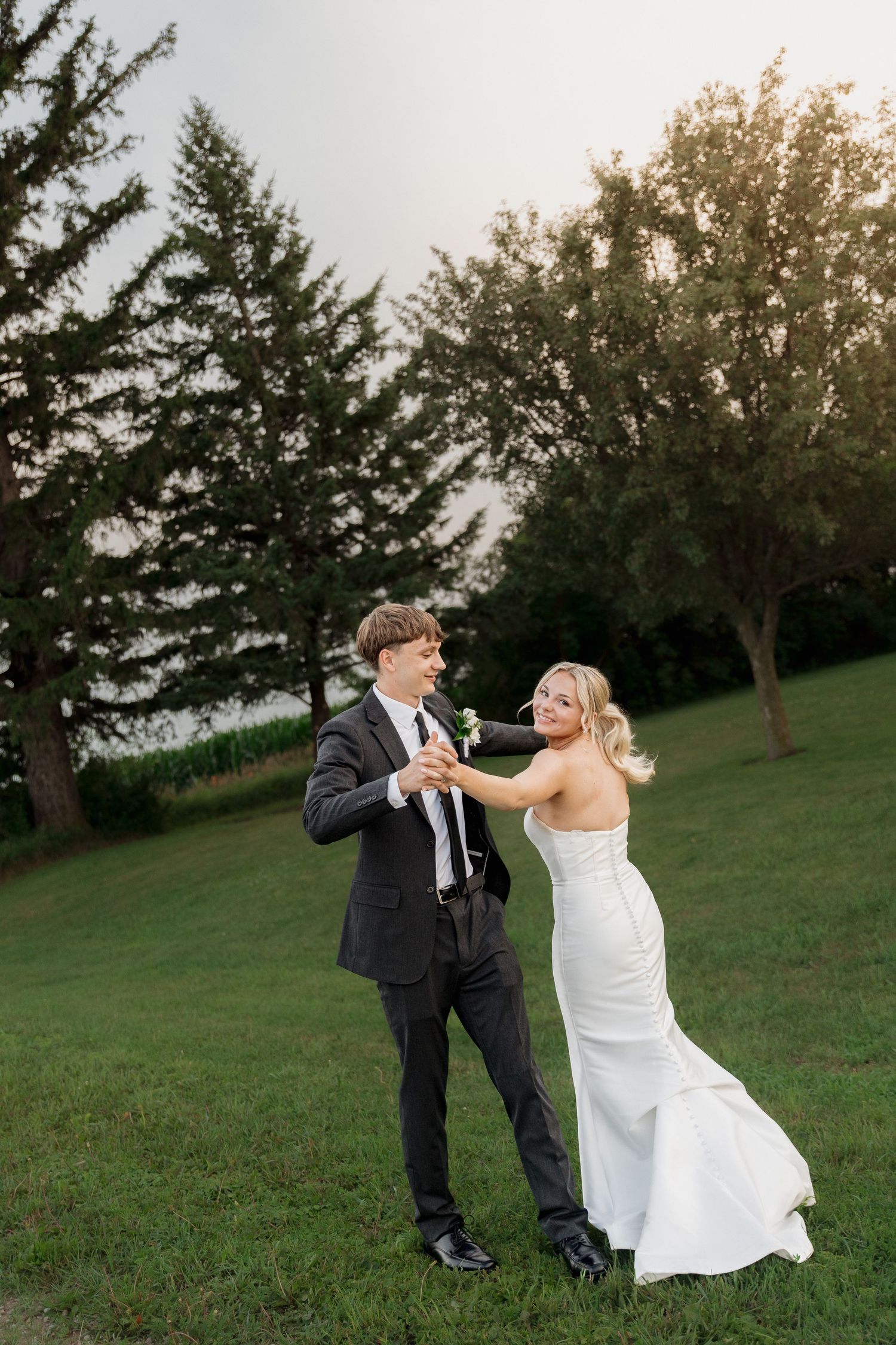 Bride and groom dancing outdoors on green grass with trees in background during golden hour wedding photos.