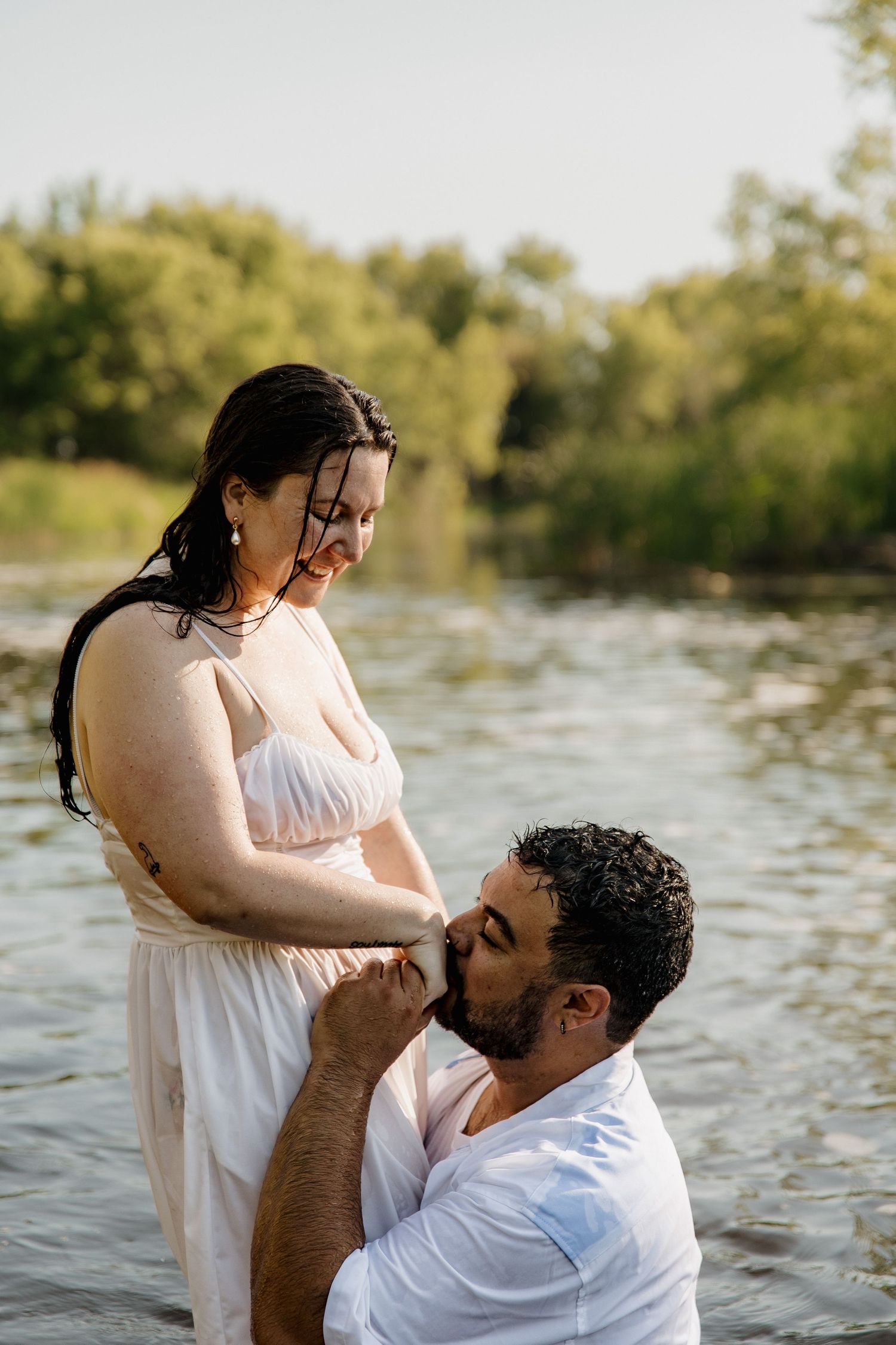 Romantic maternity photo of a couple standing in a river, man kneeling before pregnant woman in a flowing pink dress.