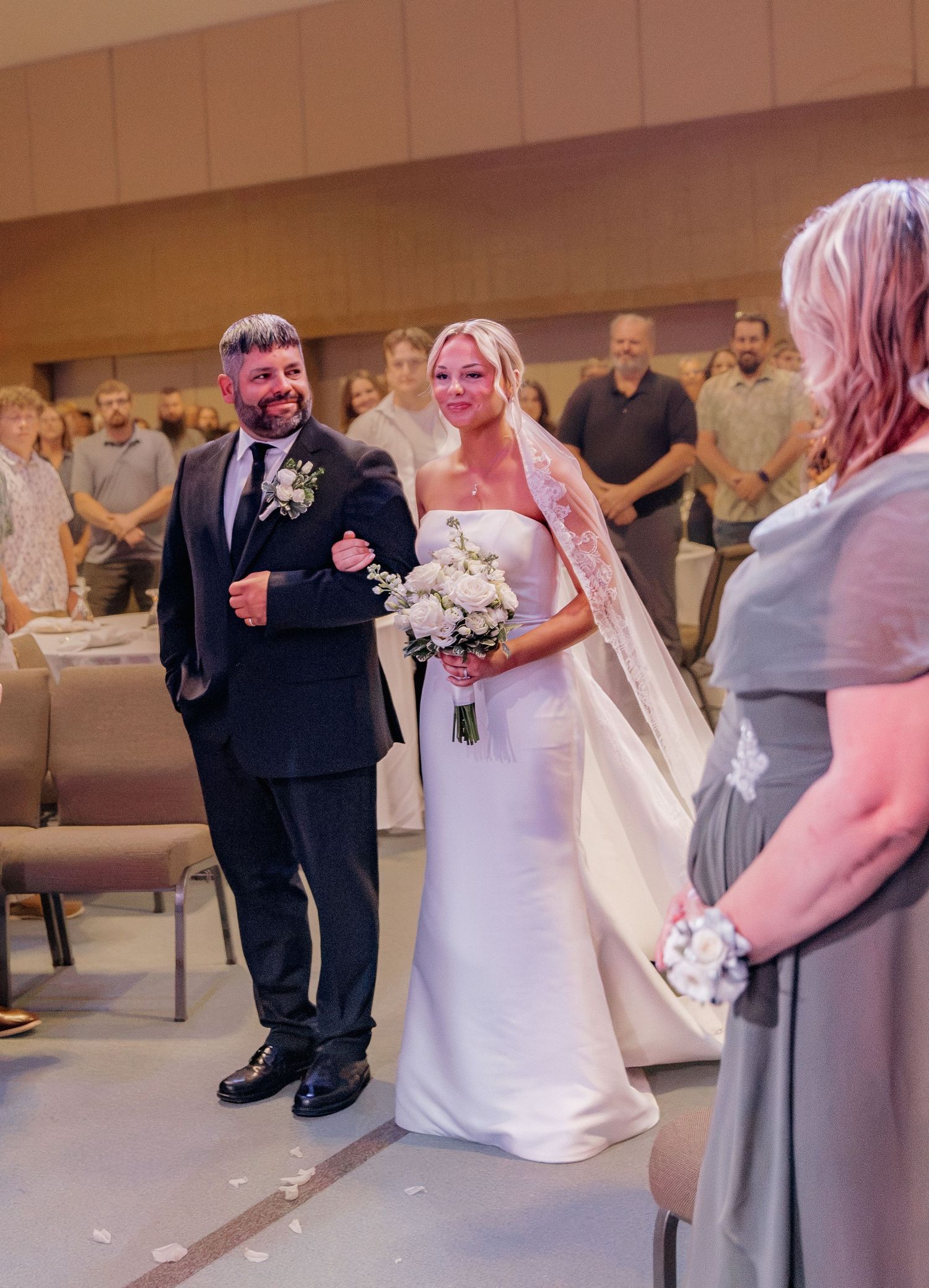 Smiling bride in white gown walks down church aisle with escort at elegant indoor wedding ceremony.