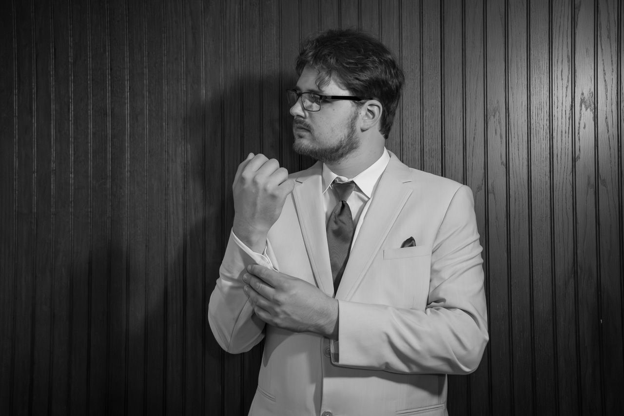 A groom in a light colored suit adjusts his tie in a dramatic black and white portrait.