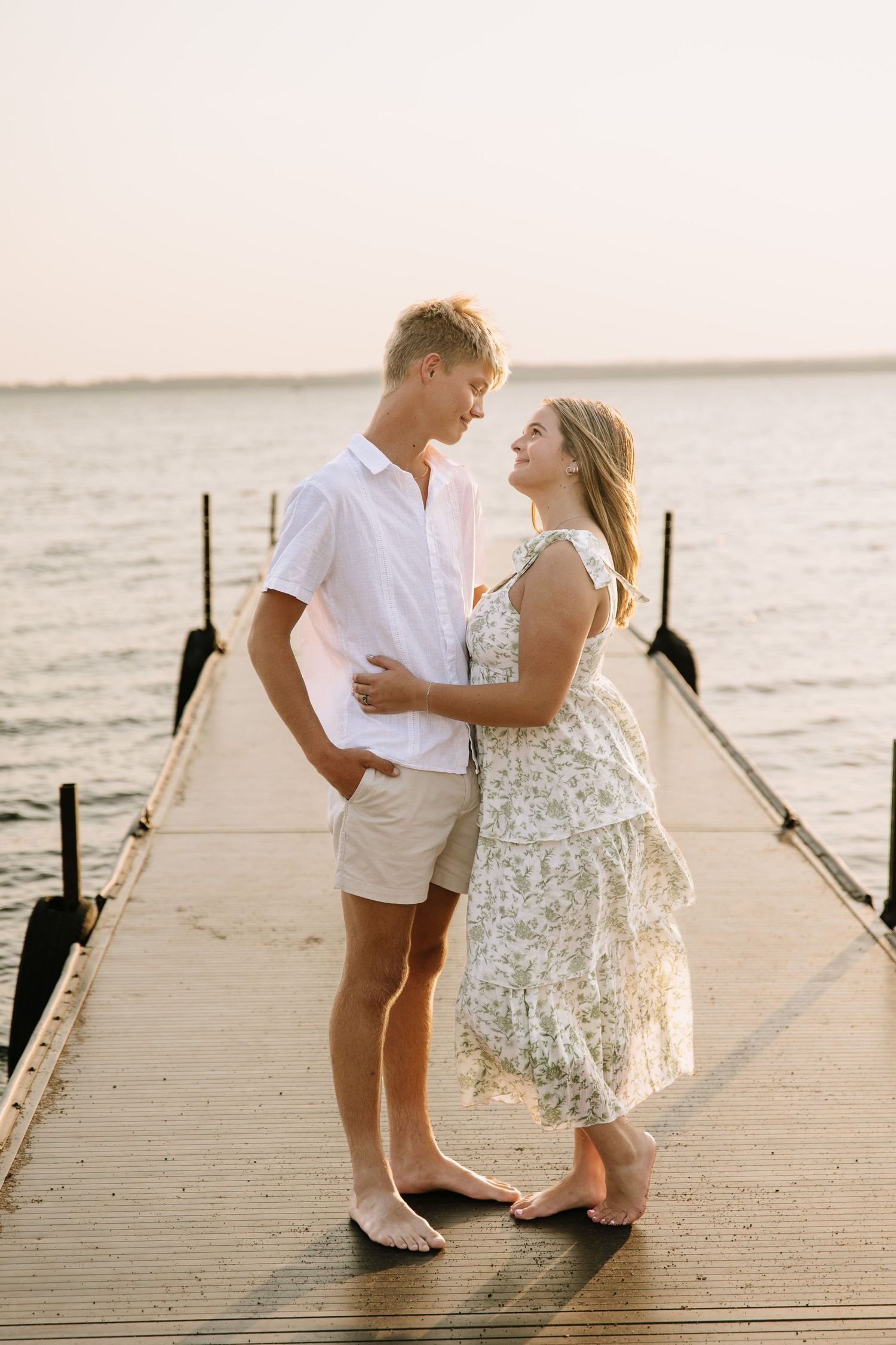 A couple embraces on a wooden dock at sunset, wearing light summer outfits, with calm ocean waters in the background.