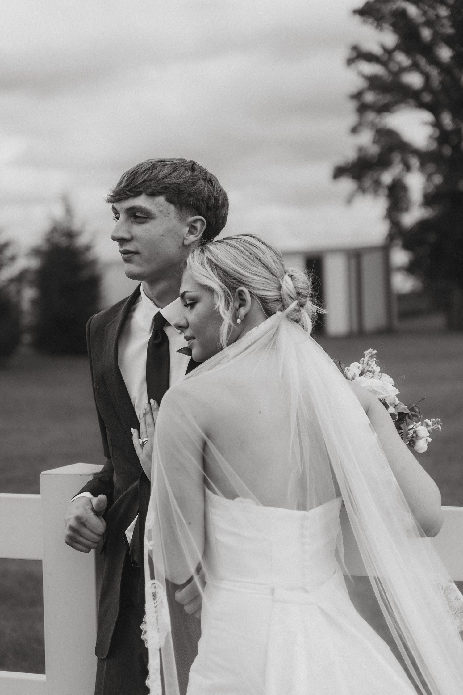 Black and white photo of bride and groom standing by white fence in outdoor setting.
