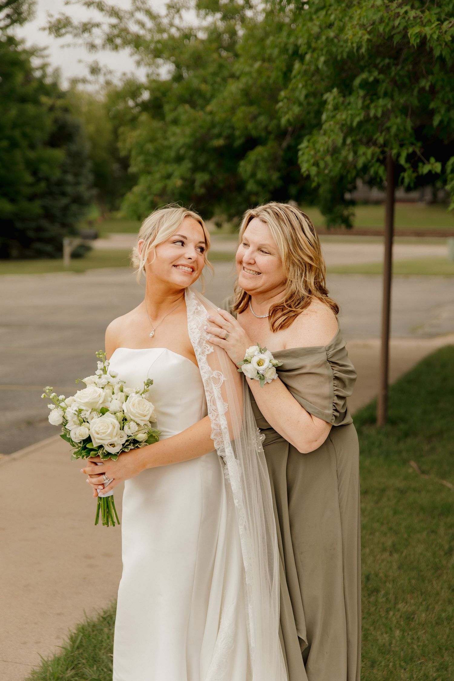Bride in strapless white gown holding white rose bouquet with bridesmaid in sage dress.
