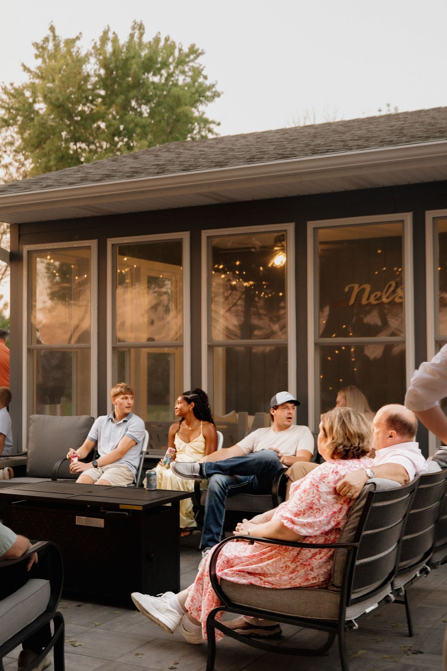 Wedding guests relaxing on patio furniture at dusk outside warmly lit venue with string lights.
