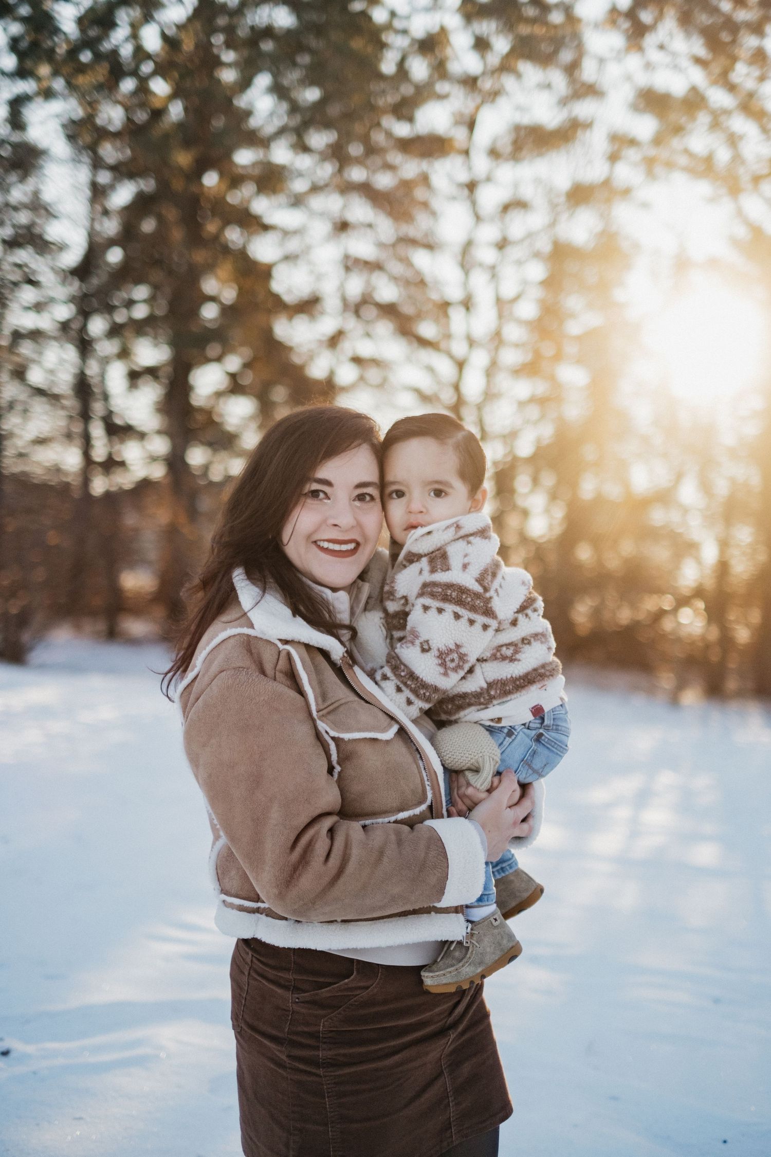 A winter portrait of a parent and child against a snowy forest backdrop at sunset.