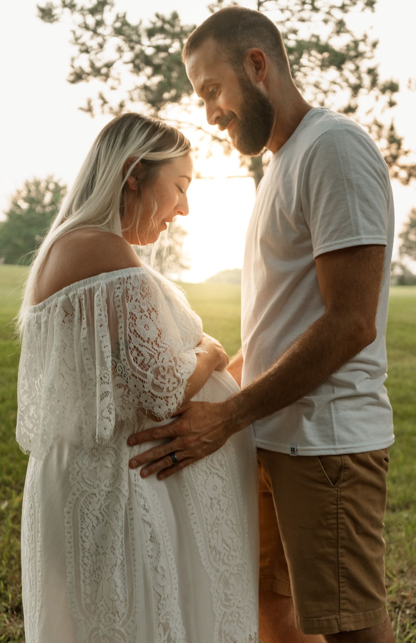 A romantic maternity photo shows a couple embracing in a sunlit field, wearing white and casual attire.