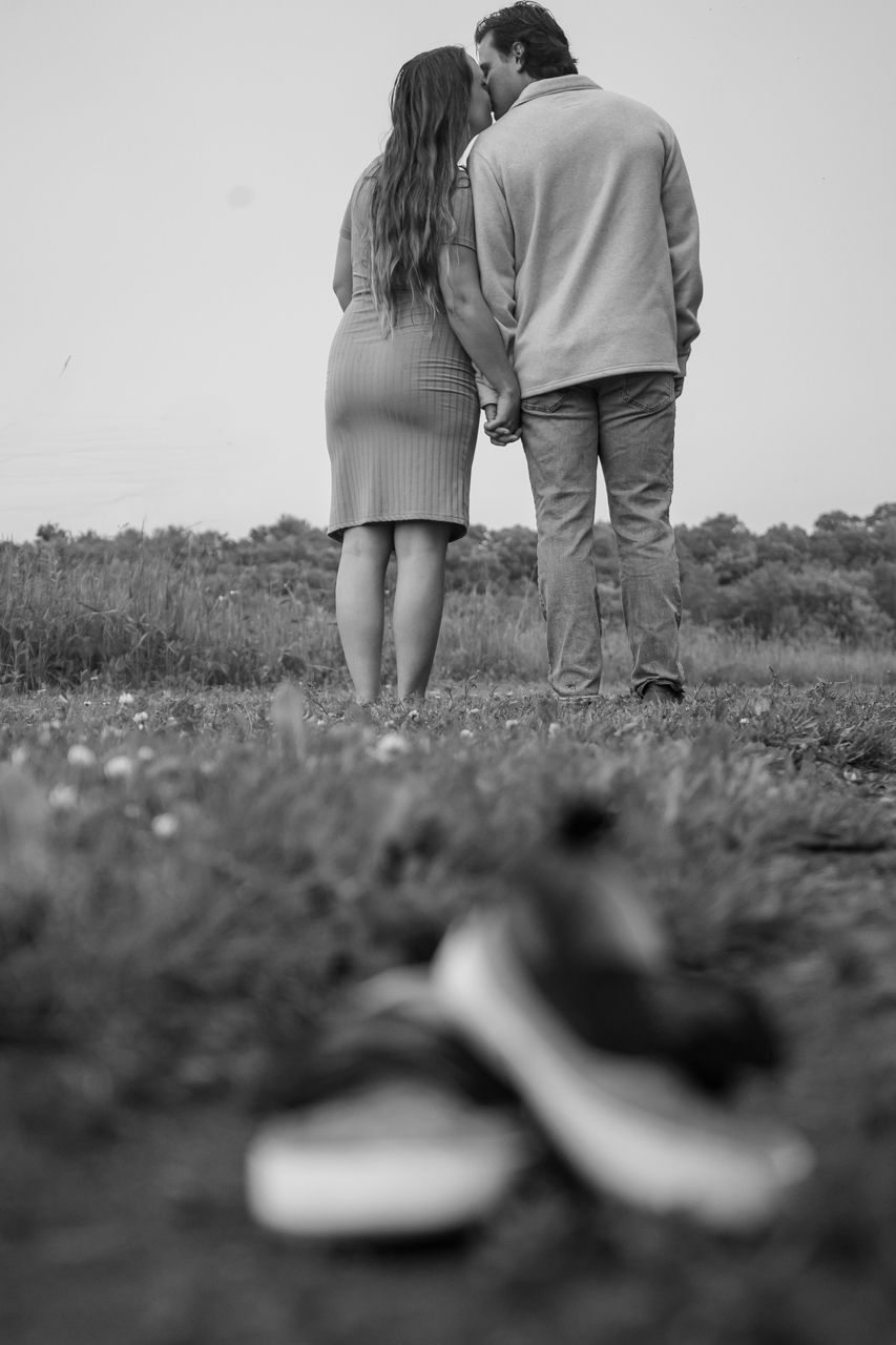 A black and white image of a couple standing in a field with baby shoes in the foreground.