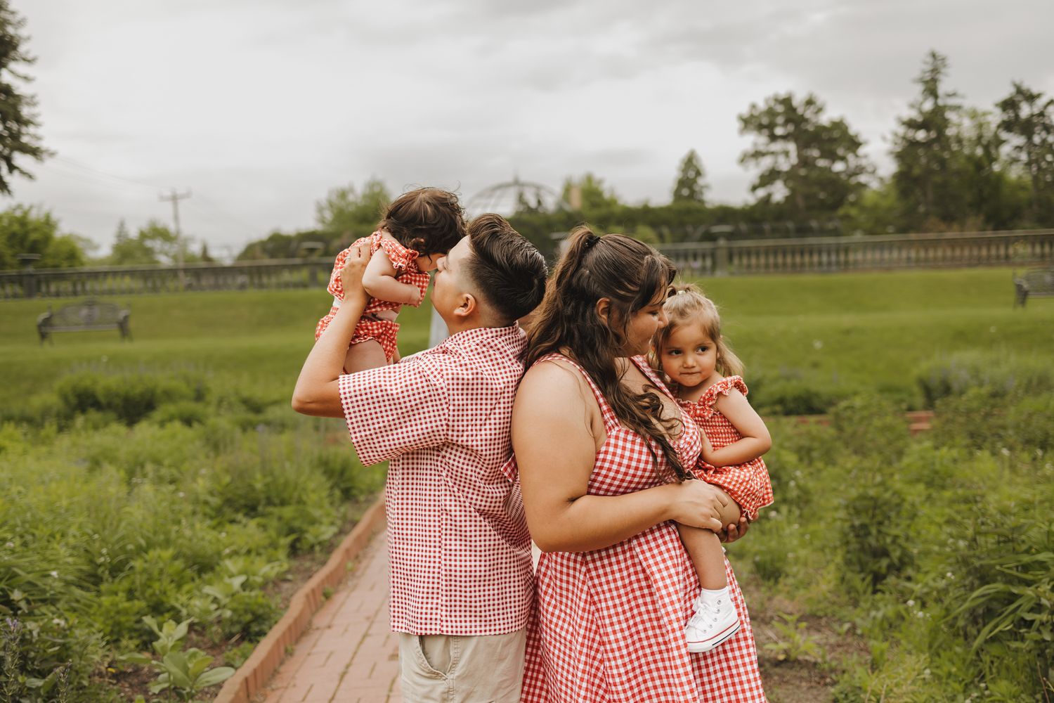 Family in matching red outfits poses outdoors on a wooden path surrounded by green fields on a cloudy day.
